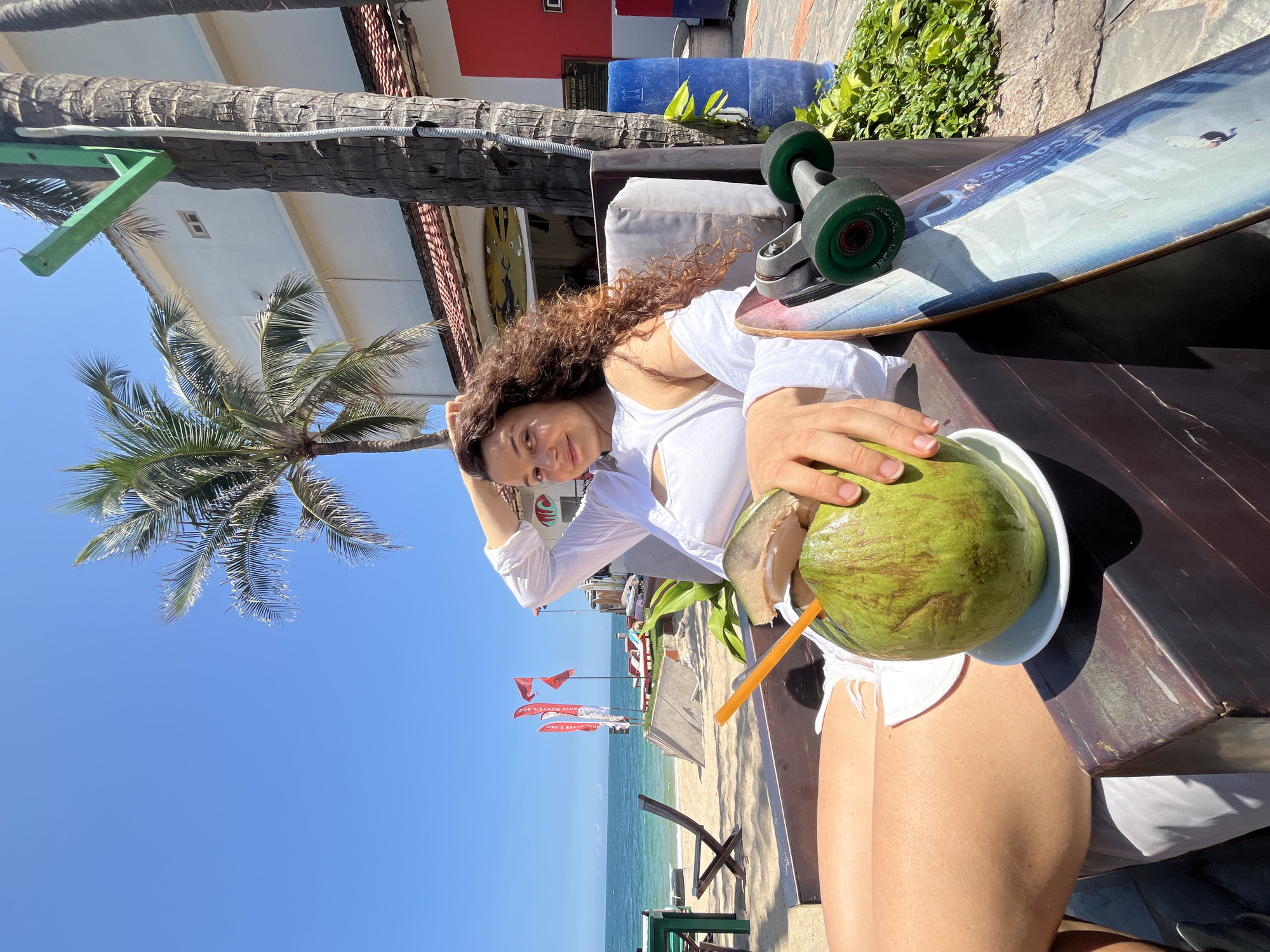 A woman with curly brown hair is sitting in a dark wooden chair on a beach in Phan Thiet, Vietnam. She is wearing a white off-the-shoulder top and white shorts. Her left hand is resting on her head, and her right hand is holding a green coconut with a straw. A skateboard is leaning against the chair to her right. In the background, the ocean and a clear blue sky are visible, along with palm trees and a building with a red and white facade. Red flags are planted in the sand on the beach.