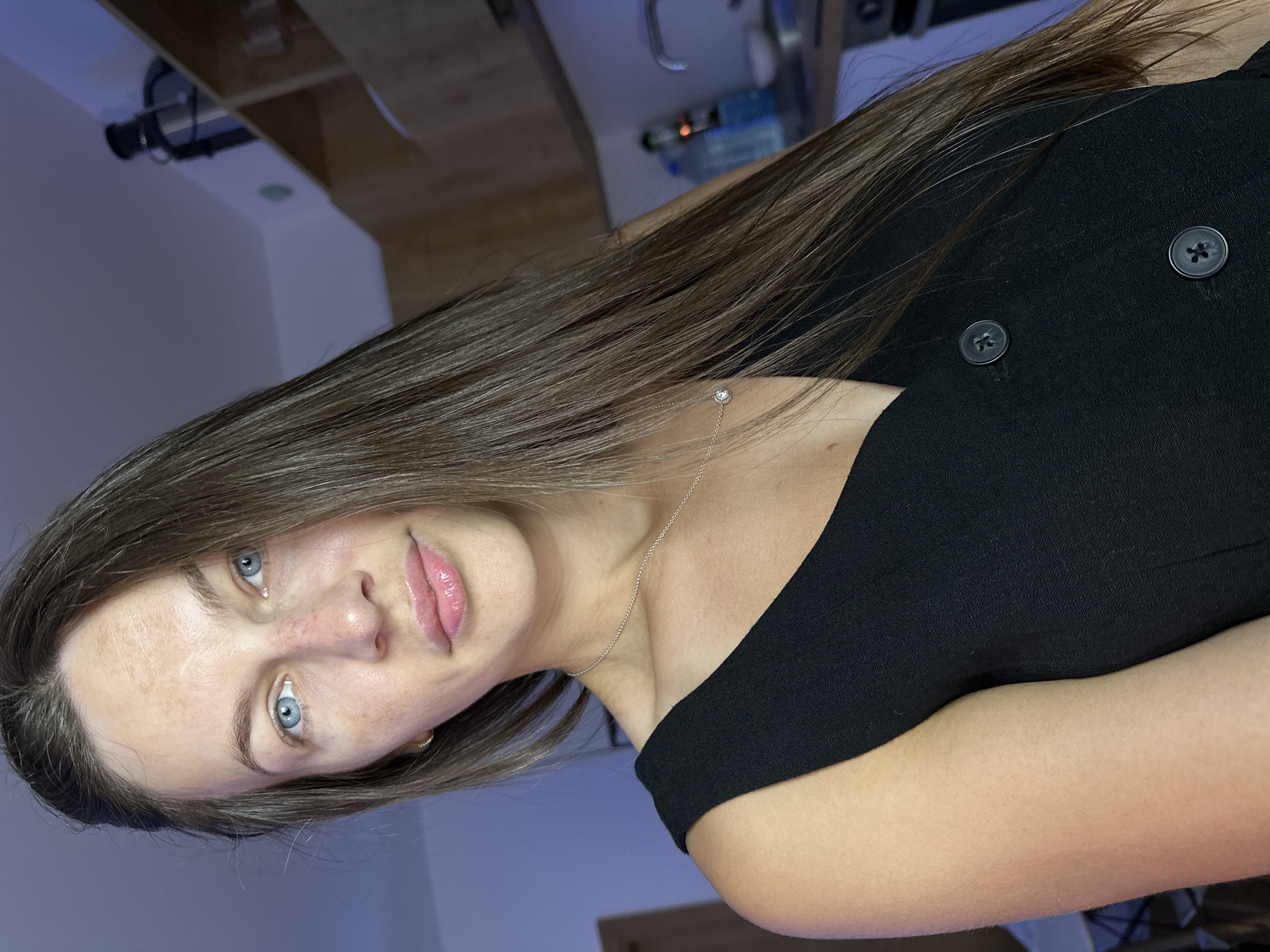 This is a portrait of a young woman with long, dark brown hair and blue eyes. She is wearing a black top with two dark buttons. A thin silver chain with a small, circular pendant rests on her collarbone. Her skin is fair with some freckles visible on her nose and cheeks. Her lips are glossy and appear to be slightly parted. The background is dimly lit and out of focus, showing hints of shelves, bottles, and what appears to be lighting fixtures. The lighting suggests an indoor setting, possibly a room or studio. The overall mood is intimate and relaxed. There is no visible text in the image.