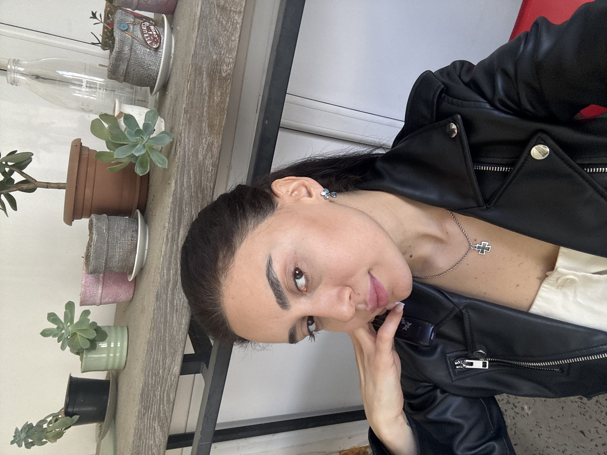 A woman is posing for a photo indoors, next to a shelf with plants in pots and a decorative glass bottle. She is wearing a black leather jacket and a light-colored top. She has dark hair pulled back, earrings, and a necklace with a cross pendant. Her hand is near her face. The plants on the shelf are various succulents and greenery. The background includes what appears to be a window and possibly a red object. The location is likely in Tbilisi, Georgia, based on the query, though the specific clues identifying it as such are not visible in this single image.