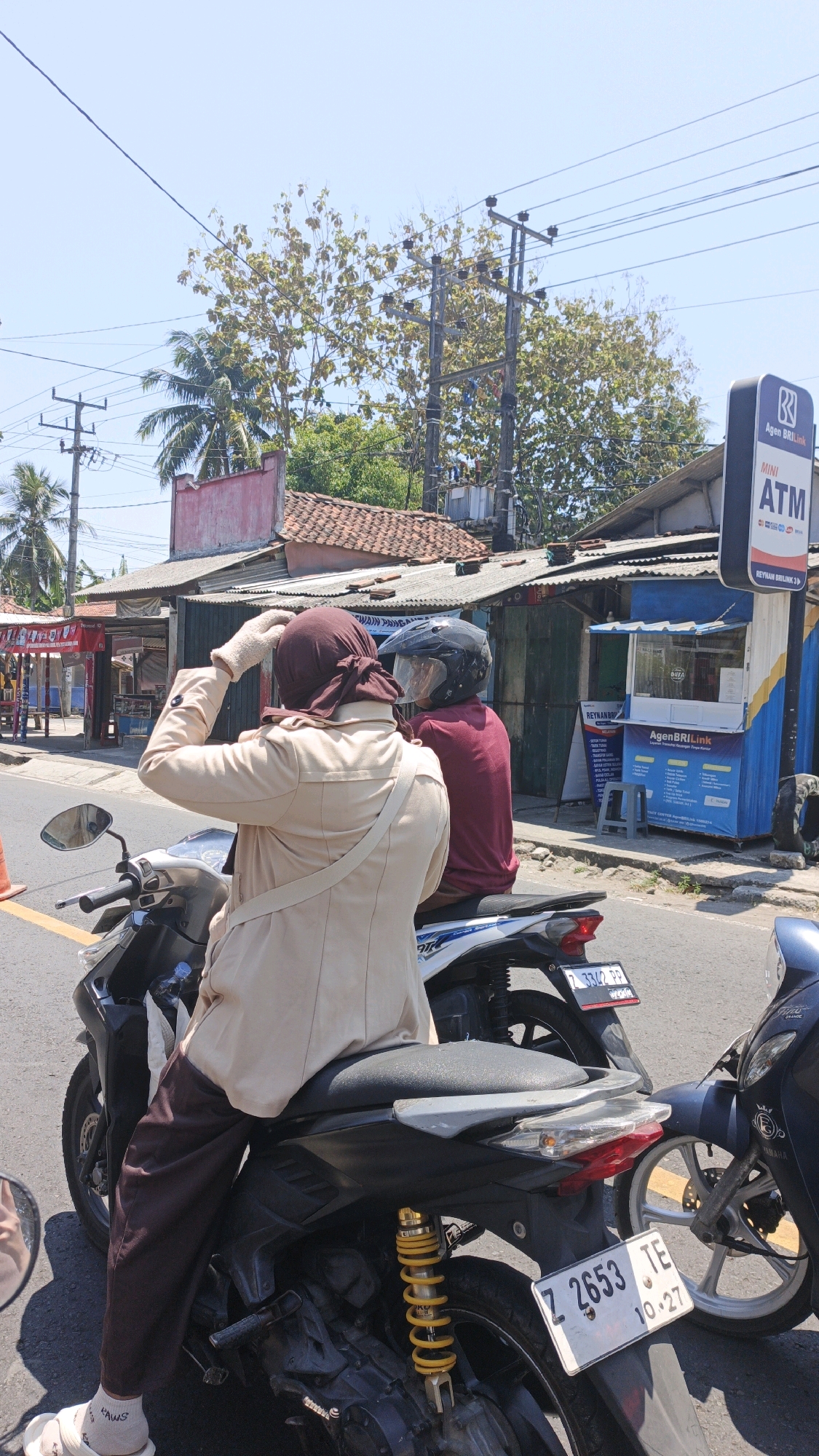 In Sukaresik, Indonesia, on a sunny day, two individuals are seated on motorcycles, with the focus on the person in the foreground. This individual, wearing a dark brown hijab and a tan jacket over brown pants, is looking to the left and adjusting their hijab. They are also wearing white socks and light-colored sandals. The person behind them is wearing a maroon shirt and a black helmet. Both are on the road, with other motorcycles parked nearby. In the background, there are buildings with corrugated metal roofs, palm trees, and utility poles with electrical wires. A blue and white ATM kiosk with "Agen BRIlink" branding is visible on the right side of the image. The license plate of the motorcycle in the foreground reads "Z 2653 TE".