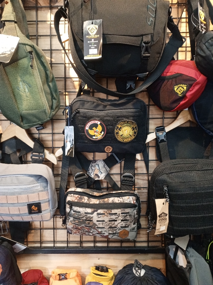This media file captures a close-up view of an indoor retail display featuring various bags and pouches, likely for outdoor or tactical use, in Baza, Myanmar. No people are visible in the image.
The scene showcases multiple bags of different sizes, colors, and designs, neatly arranged on a metal grid display wall with a wooden backdrop. In the top left, an olive green sling bag hangs, while a black messenger-style bag with a visible "OUTDOOR GEAR" tag hangs in the upper center. To its right, a red fanny pack is visible, sporting a yellow logo that appears to be a stylized 'M' and some text.
A prominent black chest rig or shoulder pouch, adorned with two circular patches, is centrally displayed. The left patch features the word "INDONESIA" above the Garuda Pancasila emblem, while the right patch shows yellow text, possibly "BUTRAM MILITER CHABAS YUSHERAC" (some letters are unclear), on a dark background. Below this, a camouflage-patterned chest rig with a MOLLE-style front is visible, also featuring a small brand logo.
To the left, a light grey pouch with horizontal webbing and a brand logo is displayed. On the right, a black textured bag with a hang tag reading "OPANAS OUTDOOR" is shown. Several other brightly colored bags are partially visible at the bottom of the frame, indicating a wider selection of products. The items are equipped with various straps, buckles, and zippers, typical of practical outdoor gear. The overall impression is that of a well-stocked store section showcasing durable and functional bags.