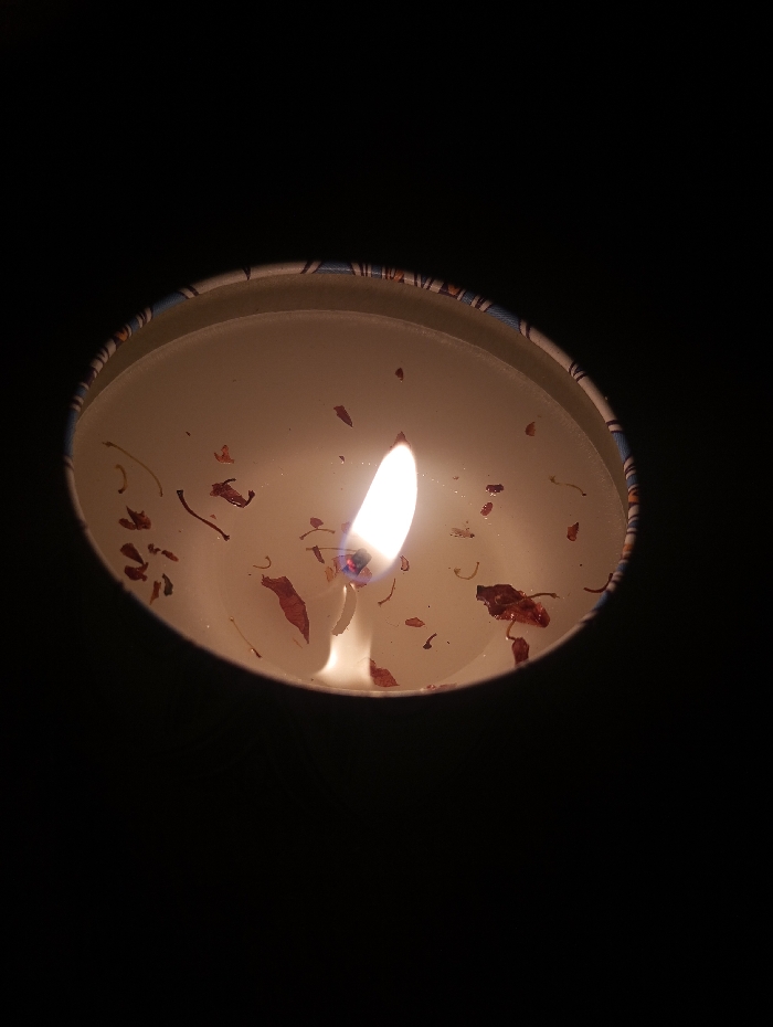 A top-down, slightly angled view depicts a lit candle enclosed within a circular vessel. The candle consists of light-colored wax, with a central, elongated flame emitting a bright yellow-white light. The flame illuminates a molten pool of wax, where numerous small, irregularly shaped, dark reddish-brown organic fragments are suspended. The outer rim of the vessel features a narrow band with a multi-colored, patterned design. The immediate surrounding environment is uniformly black, indicating the candle is the primary light source. This scene is located in Guacara, Venezuela.