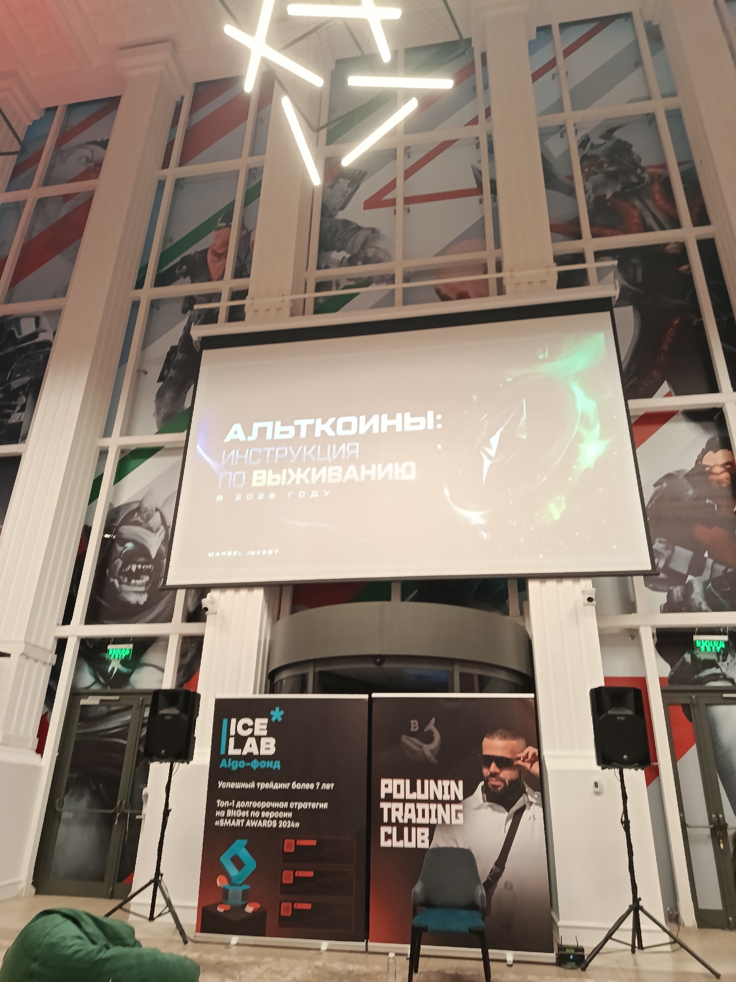 The image shows a presentation setting, likely at a conference or seminar, with a projector screen displaying the title "Альткоины: Инструкция по выживанию" (Altcoins: Survival Guide) in Russian.
Below the screen is a stage area with two banners. One banner is for "ICE LAB Algo-фонд," advertising successful trading for over 7 years and the top long-term strategy on BitGet according to "SMART AWARDS 2024." The other banner displays "POLUNIN TRADING CLUB" along with an image of a man wearing sunglasses.
A chair sits in front of the Polunin Trading Club banner. Speakers on stands are placed on either side of the stage area.
The setting appears to be indoors, with large windows behind the presentation area, showcasing artwork or designs in each pane. The lighting is a combination of natural light from the windows and artificial light from a modern chandelier. The environment is possibly a conference hall or event space within a larger building. The context seems to be related to cryptocurrency, finance, and trading strategies. The image provides no indication that the location is in Moscow, Russia.