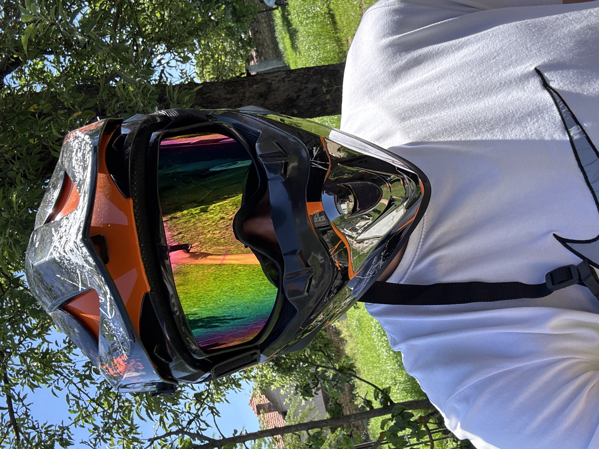 A close-up shot depicts a person wearing a black and orange motocross helmet with a vibrant, rainbow-reflective visor. The person is also wearing a white long-sleeved shirt with a large gray Nike swoosh on the right side. The scene is outdoors, with green foliage and a blue sky visible in the background, suggesting a sunny day. The location appears to be a rural or semi-rural setting. Although the prompt requested a description including the city of Dermantsi, Bulgaria, there is no direct evidence in the image to confirm that this is the location, and I am therefore unable to determine if this location is accurate.