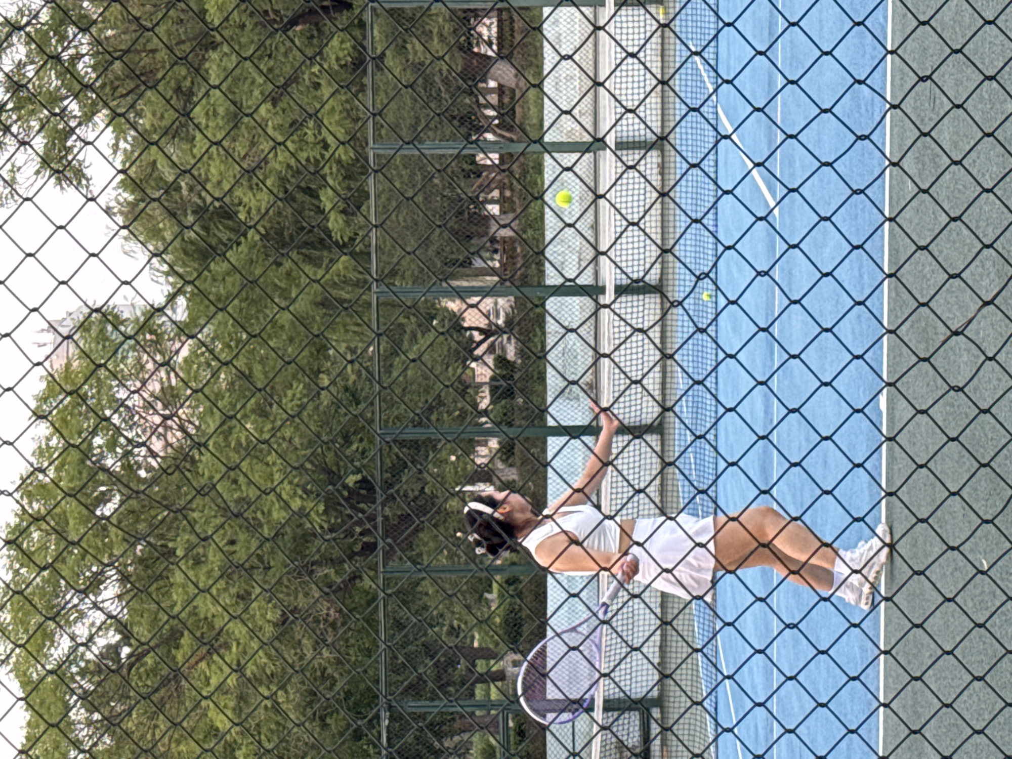 A young woman wearing a white tennis outfit and a white headband is playing tennis on a blue hard court. She is in mid-swing, about to hit a yellow tennis ball. A tennis racket is visible in her hand. The court is enclosed by a green chain-link fence, which is in the foreground of the image. Beyond the fence, trees and buildings are visible. The image appears to have been taken in Linghe, China.