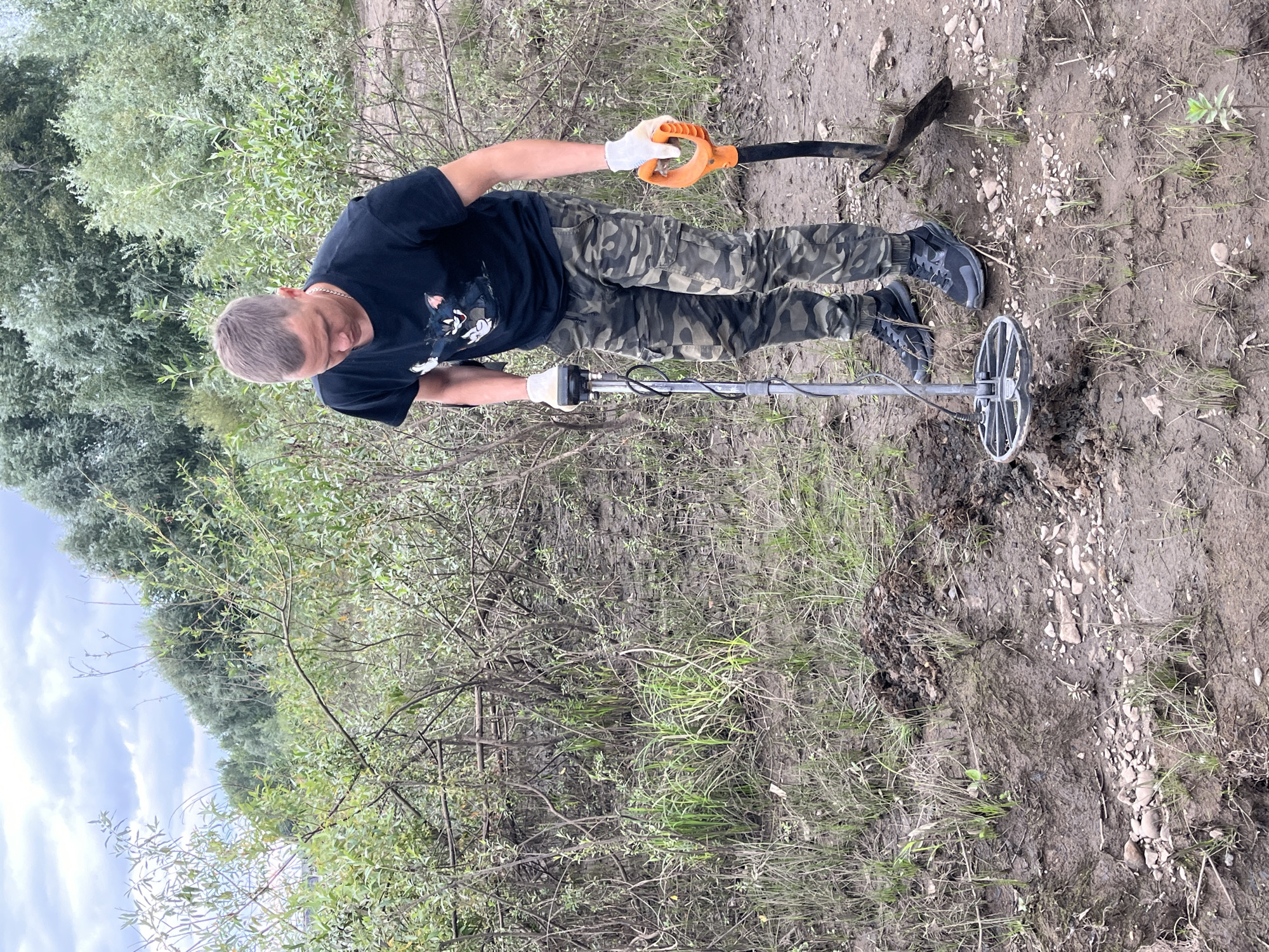 A man wearing a black t-shirt, camouflage pants, white gloves, and black sneakers is metal detecting in a muddy, vegetated area. He is holding a metal detector with a circular coil near the ground, and to his right, a shovel with an orange handle is stuck in the dirt. The background features dense green foliage and a cloudy sky. The location is likely the banks of the Kama River in Chaikovsky, Russia, a region known for its industrial history and potential for metal object discovery. The muddy terrain suggests recent rainfall or proximity to a water source. The man's attire and equipment indicate an activity related to treasure hunting or archaeological surveying.