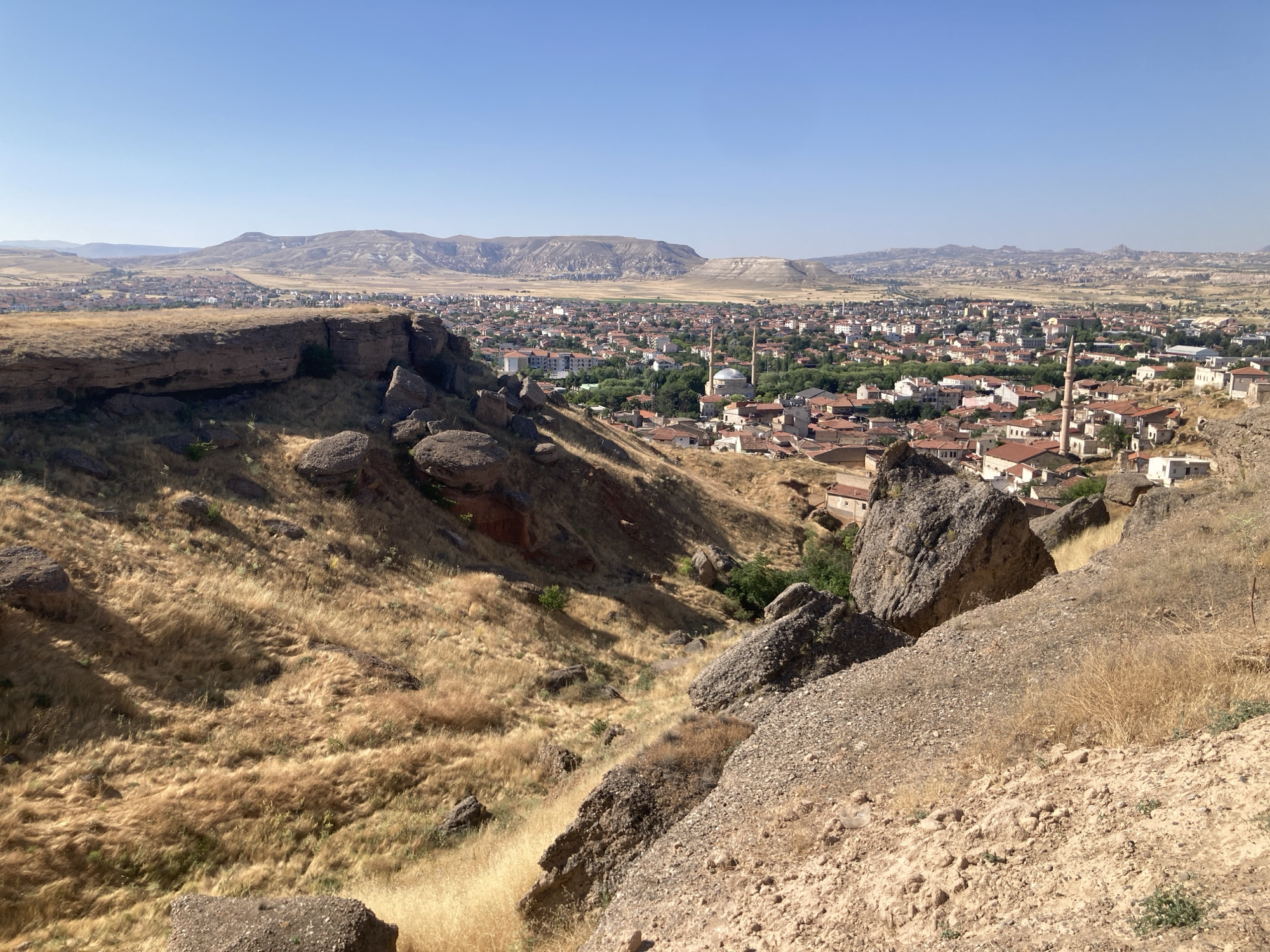 This is a wide-angle, landscape photograph taken from an elevated vantage point overlooking the city of Avanos İlçe Merkezi, Turkey, on a clear, sunny day. The foreground is dominated by a rugged, rocky slope covered in dry, golden grass, indicative of a hot, arid climate. Large boulders are scattered across this terrain.
Beyond the immediate foreground, the terrain slopes down towards the city, revealing a dense urban area with predominantly red-tiled roofs. Numerous buildings, varying in size and style, are packed closely together. Key architectural features visible include several minarets, suggesting the presence of mosques, and at least one building with a dome. Greenery, in the form of trees and shrubs, is interspersed throughout the city, providing pockets of contrast against the predominantly reddish-brown hues of the buildings and landscape.
In the distance, a range of hazy, mountainous terrain stretches across the horizon, its undulating forms softened by atmospheric perspective. The sky is a clear, unblemished blue. There are no people or animals visible in the scene. The overall impression is one of a historically rich, densely populated settlement nestled within a dramatic, natural landscape.