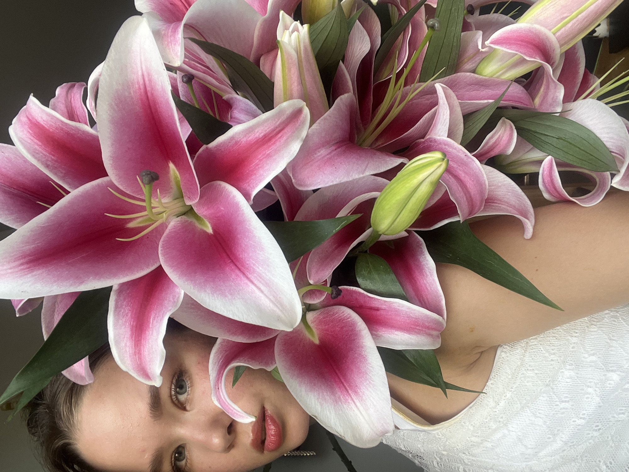 A young woman lies on her side with a bouquet of pink and white lilies arranged around her head. Her face is visible at the bottom of the frame, while the lilies dominate the upper portion. She wears a white, lacy garment. The background appears to be a muted gray. The image does not appear to be taken in the city of Nam Natural Heritage Area Trang, Vietnam, as there are no elements in the frame suggesting an outdoor or Vietnamese location.