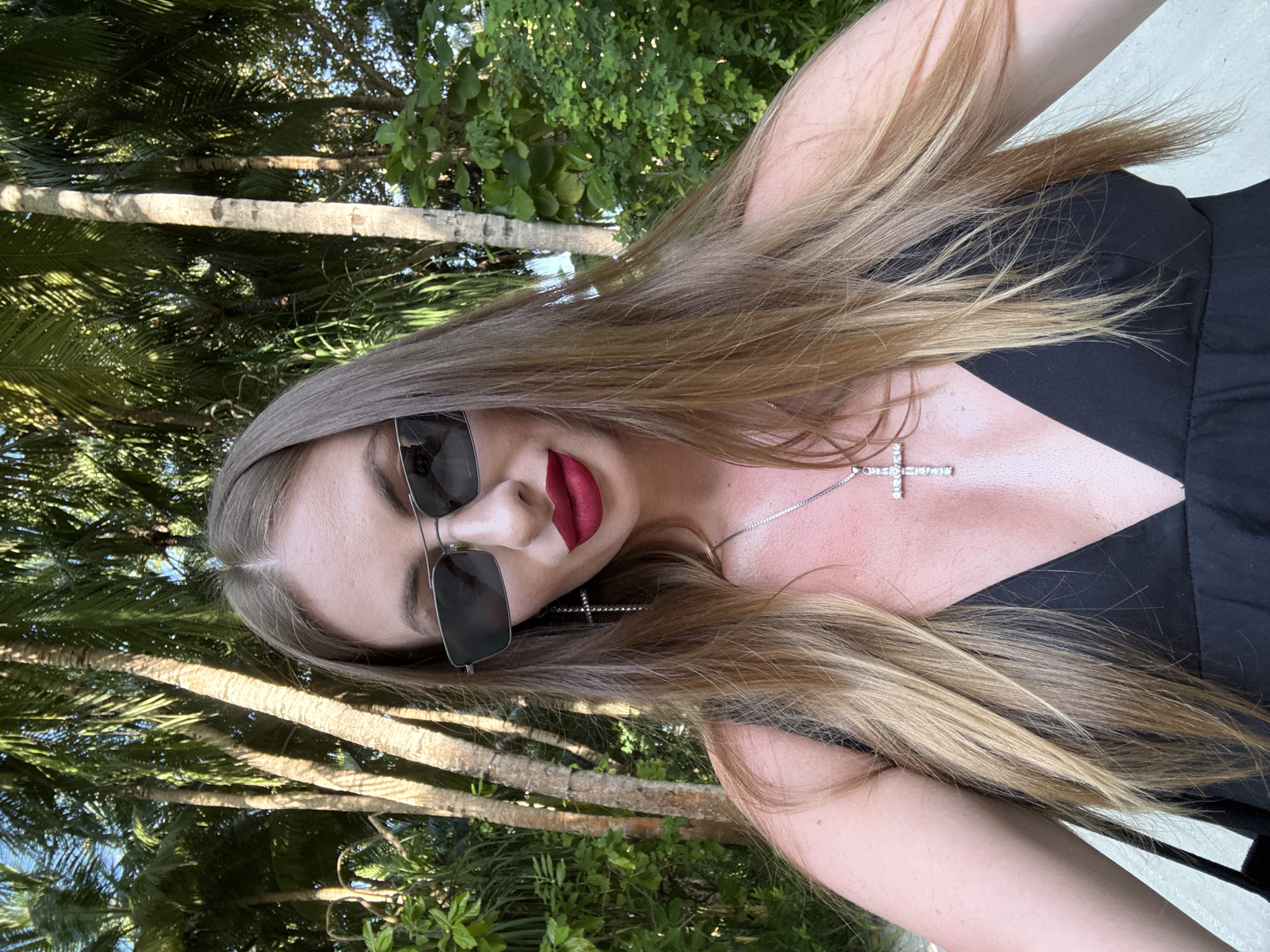 The image shows a close-up, portrait-style selfie of a woman with long, blonde hair. She is wearing sunglasses and has red lipstick. A silver necklace with a cross pendant is visible around her neck. She is wearing a black top, and her skin appears slightly shiny, suggesting she might be in a warm, possibly humid environment.
The background consists of lush green foliage and palm trees, with some dappled sunlight filtering through. This suggests an outdoor setting, likely in a tropical or subtropical location. The presence of palm trees and the mention of "Maldives" as a location cue strongly point towards a beach or island setting. The lighting and the bright green of the leaves suggest it is daytime, possibly late morning or afternoon.
The woman's pose and direct gaze into the camera are typical of a selfie. Her facial expression, with her lips slightly parted and makeup applied, could be interpreted as playful or confident. There is no visible text in the image. The overall impression is one of a casual, vacation-like moment captured outdoors in a tropical environment.