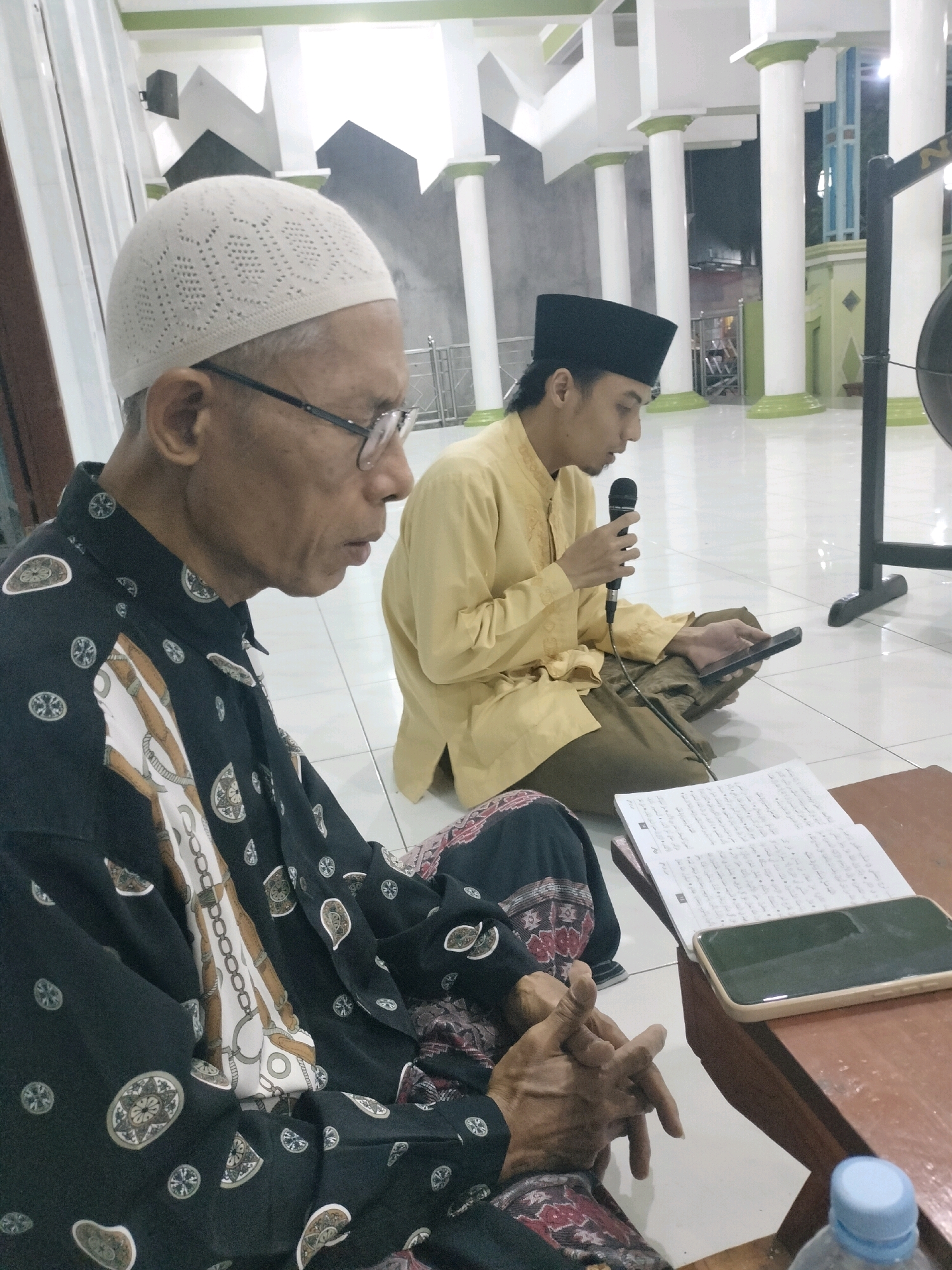 In Wedung, Indonesia, two men are seated on the floor of a mosque. The man on the left, wearing a white skullcap and a black patterned shirt, is looking to his right with his hands clasped. The man on the right, wearing a black peci and a yellow tunic, is holding a microphone in his right hand and a smartphone in his left hand. He is looking down at the smartphone, and an open book is placed on a wooden table in front of him. The mosque interior features white tiled floors and prominent white pillars with green accents. The scene is lit by artificial lights, casting a warm glow on the subjects and the surrounding architecture.