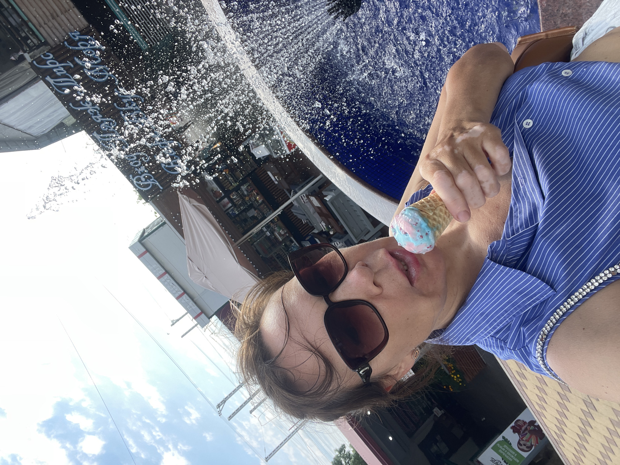 A woman is holding an ice cream cone and eating it in front of a fountain. The ice cream cone has a waffle cone and the ice cream is light blue and pink. The woman is wearing sunglasses and a blue and white striped shirt. She has brown hair and appears to be in a public outdoor space, possibly a boardwalk or plaza, with a fountain spraying water in the background. The scene is brightly lit, suggesting it is daytime, with a clear blue sky. In the background, there are buildings with awnings and some signage, hinting at a commercial or tourist area. One sign is visible in the foreground and appears to be in Cyrillic script, with the word "МОРОЖЕНОЕ" (ice cream) visible, confirming the presence of an ice cream vendor. The overall impression is one of a relaxed, enjoyable summer day.