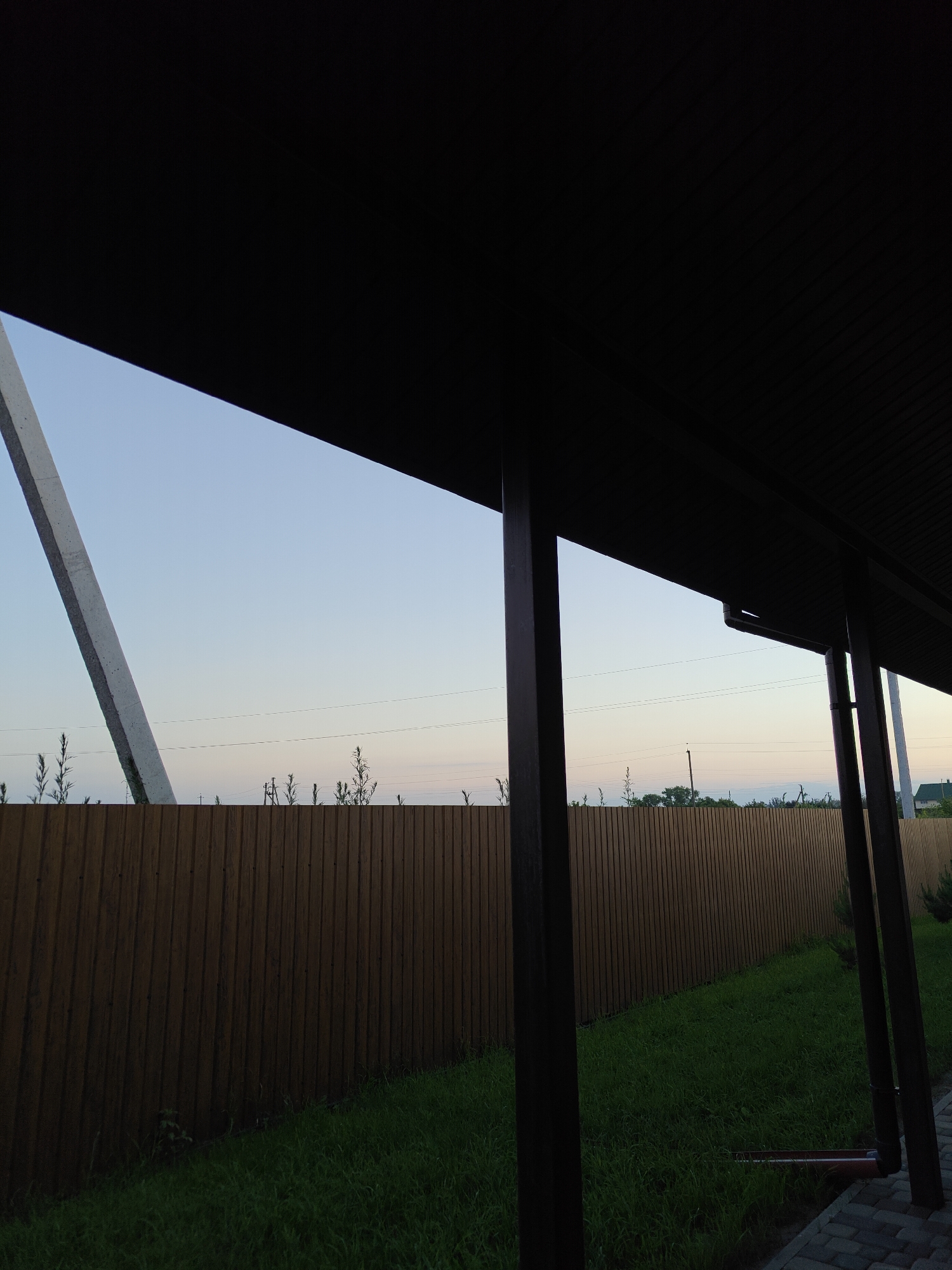 The image depicts an outdoor scene with a prominent dark brown roof structure in the foreground, casting a shadow over the view. Beyond the roof, a wooden fence runs horizontally across the frame, separating the property from the surrounding environment. The sky is a clear, pale blue, suggesting it is either early morning or late evening. A tall, light-colored concrete structure is visible to the left, leaning against the sky. Small bushes and some electrical wires are seen above the fence line. The ground is covered in green grass, with some paving stones visible in the lower right corner. The scene appears to be a residential area.