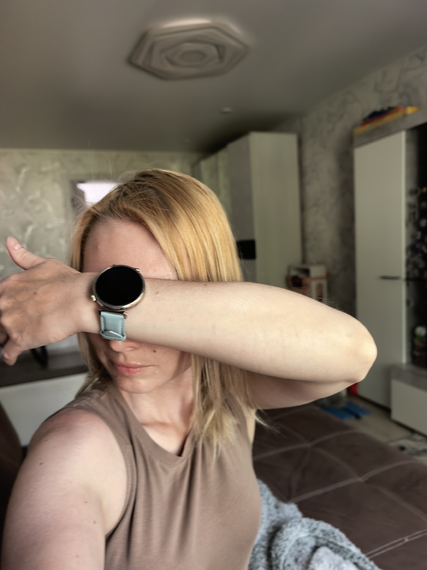 A woman is posing indoors, obscuring her face with her left forearm and hand, on which she is wearing a smartwatch. The watch has a round face and a light blue strap. She is wearing a sleeveless tan top. The background includes furniture such as a brown sofa and white cabinets. The location is Sterlitamak, Russia.