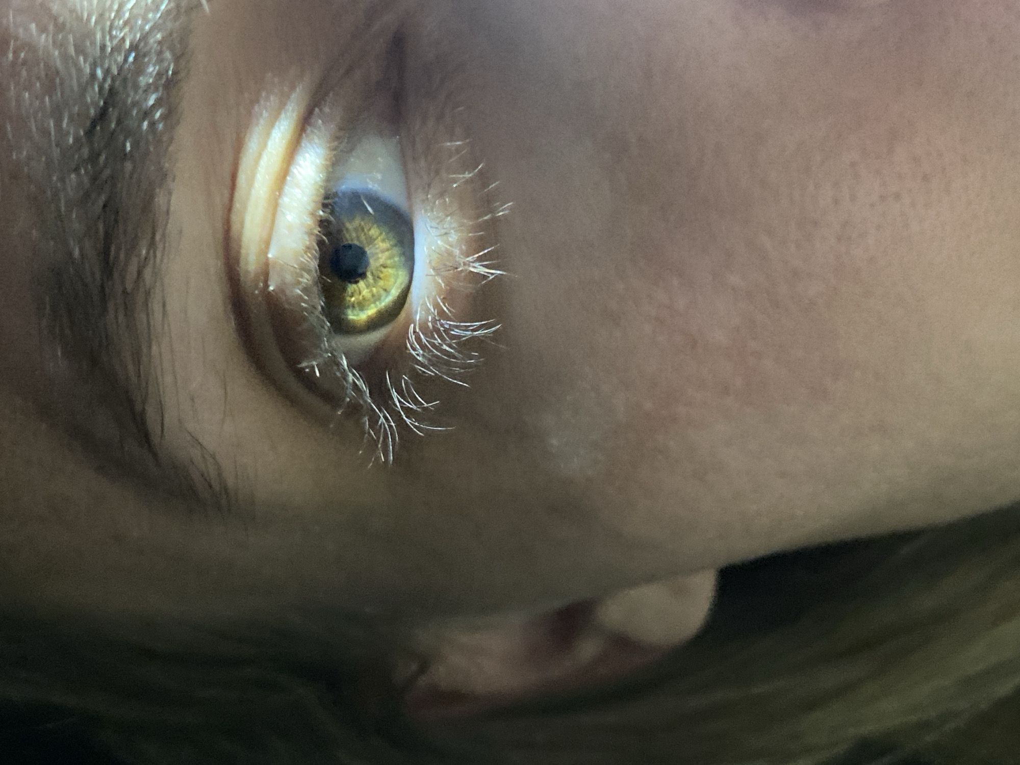 A close-up, angled view shows a human eye with a hazel iris. The eyelashes are light in color. The surrounding skin is pale and textured. Part of an ear is visible in the lower center of the image. Hair is visible along the bottom of the frame. The location is not specified. There is no description or visual evidence indicating the city of Chaikovsky, Russia.