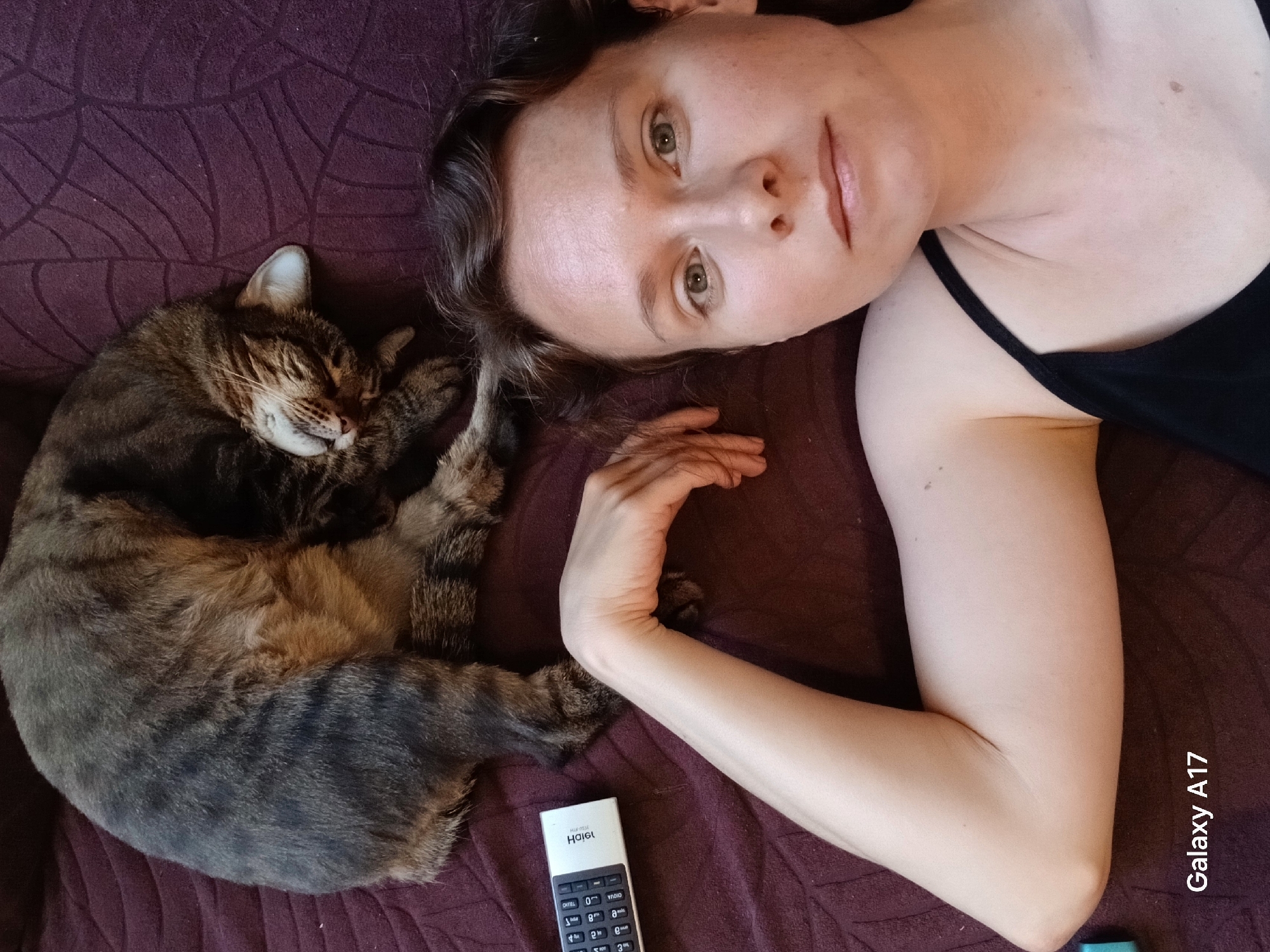 A woman lies on a purple patterned surface, with her head near a sleeping tabby cat. Her right arm is extended, with a remote control resting near her hand. The woman is wearing a black tank top. The words "Galaxy A17" are visible in the bottom right corner of the image, likely indicating the device used to take the photo.
I am unable to provide a location in the city of Yekaterinburg, Russia from this image. There is no data to support this claim.