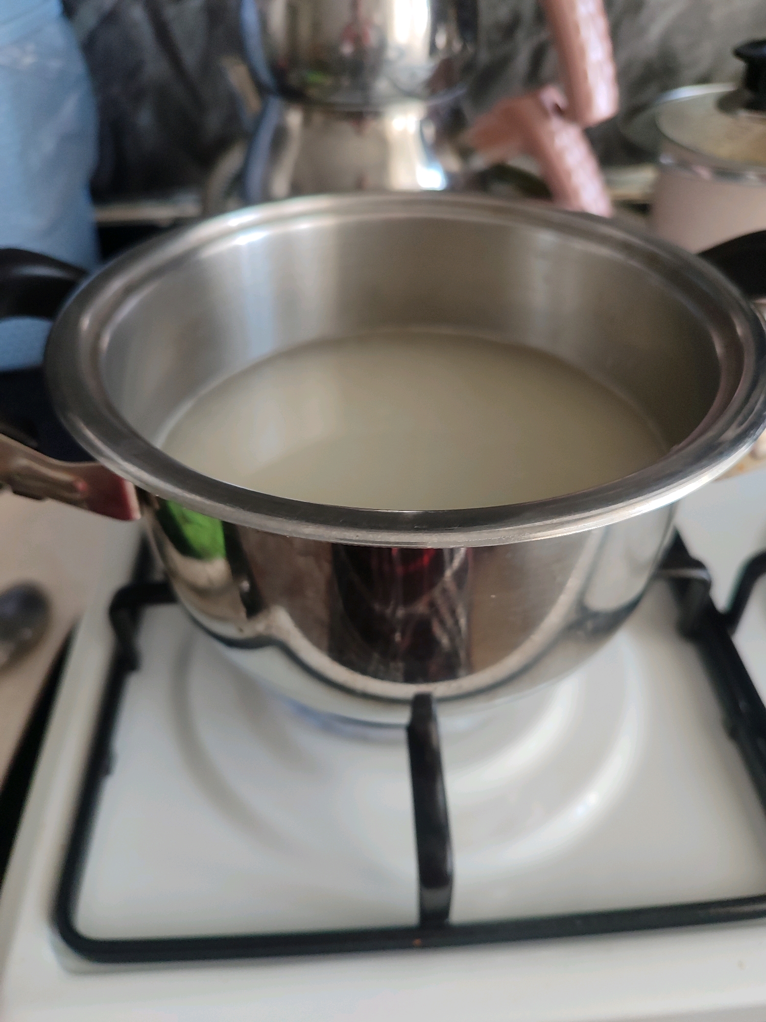 A close-up, slightly angled shot shows a stainless steel pot filled with a pale liquid, likely water or broth, sitting on a white gas stove. The pot has black handles and reflects the surrounding environment. The stove has a white enamel surface with black grates and knobs. In the background, out of focus, are various kitchen items including what appears to be a kettle or another pot, and possibly a roll of paper towels. The scene suggests a domestic setting, likely a kitchen, in Gümüşova, Turkey.