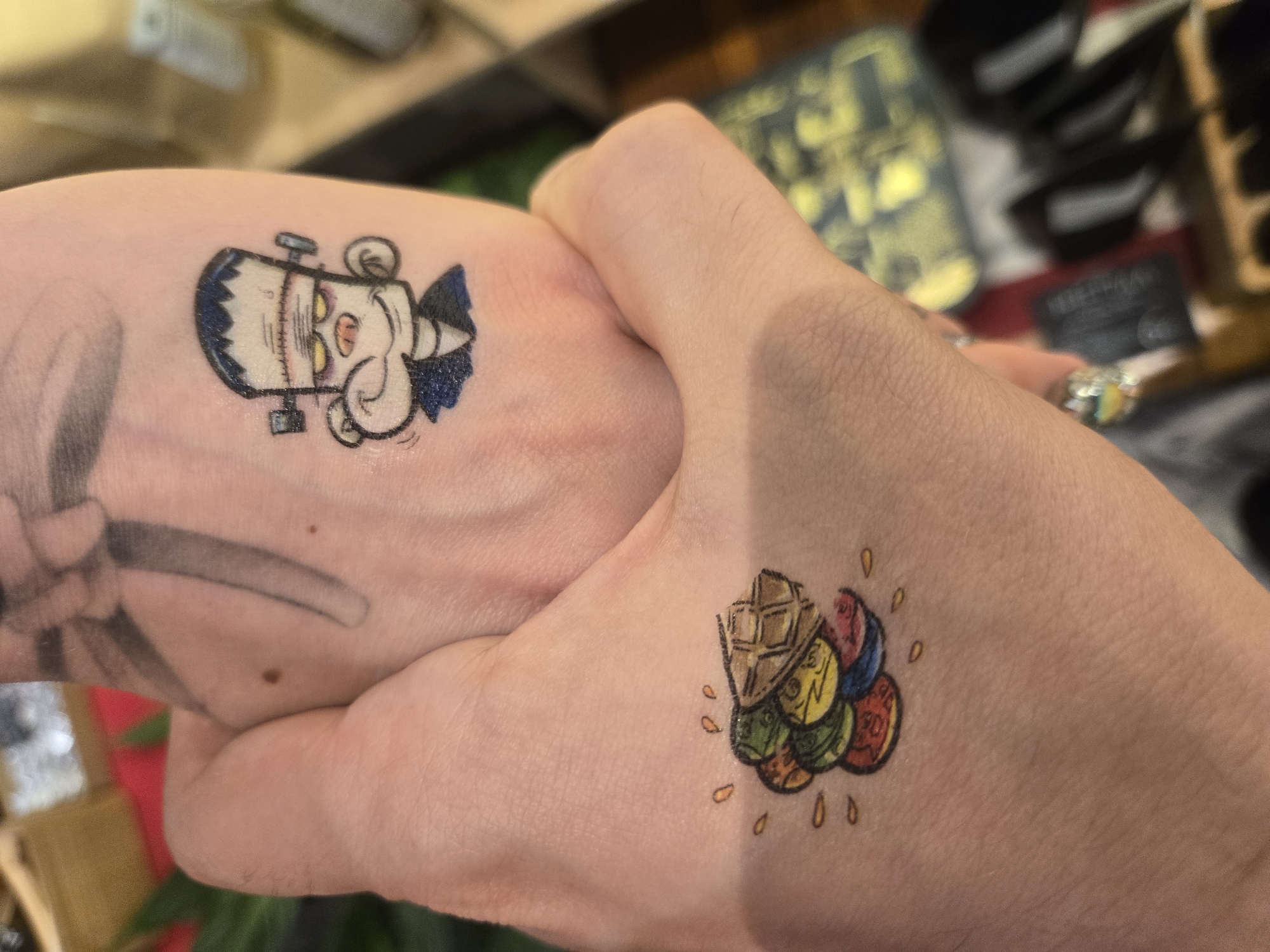 The image shows two hands clasped together, each with a tattoo. The hand on the left has a tattoo of a cartoonish Frankenstein's monster head, with exaggerated features and a cheerful expression. The hand on the right has a tattoo of what appears to be a waffle cone with multiple scoops of brightly colored ice cream. The background is out of focus, but hints of objects and possibly greenery are visible. The location of Budva, Montenegro, cannot be determined from this image.