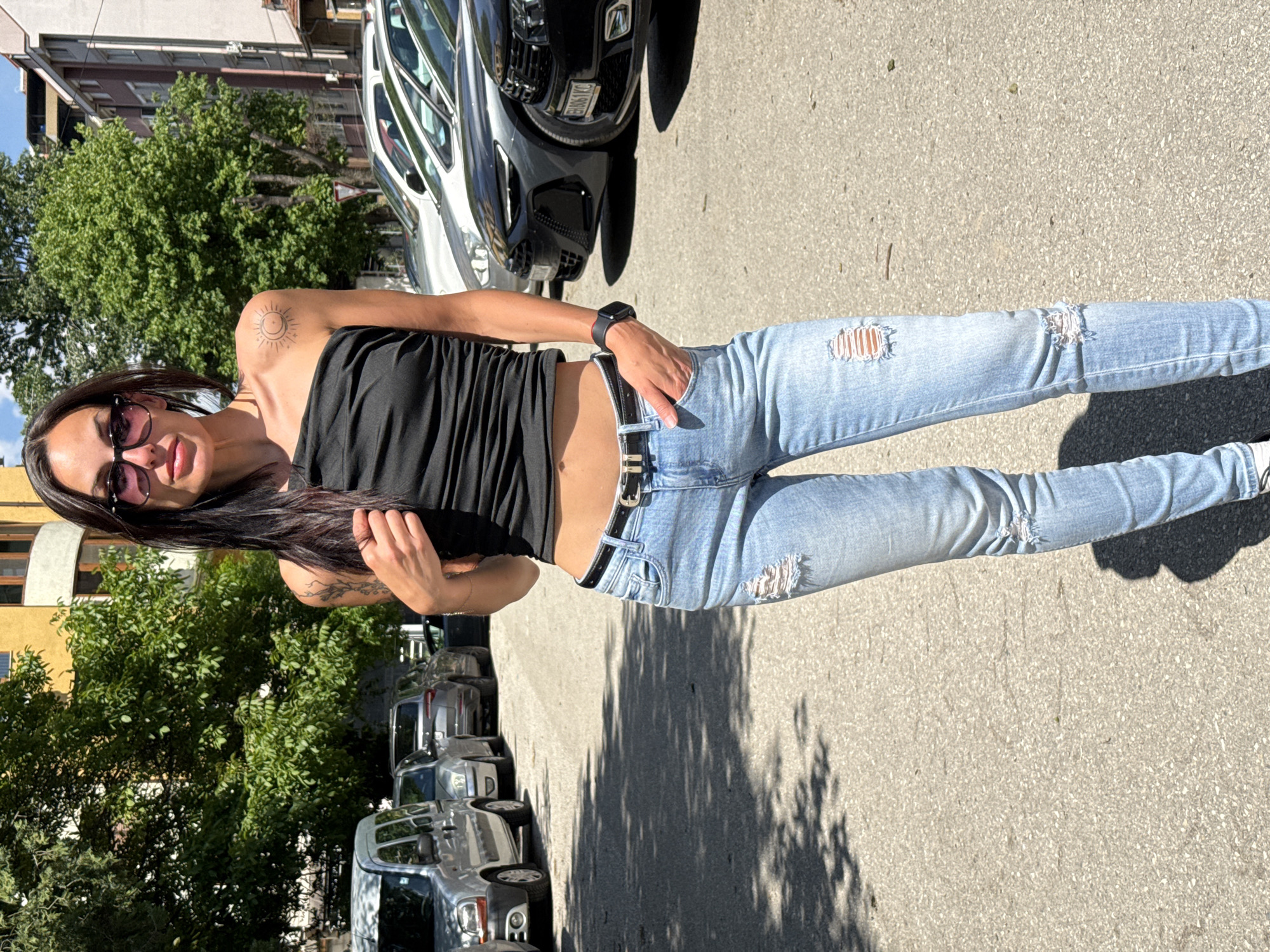A woman stands on a paved street in Pleven, Bulgaria. She wears a black strapless top, light blue ripped jeans, and sunglasses. She has long dark hair and tattoos on her arm and shoulder. Her left hand is in her pocket. Cars are parked along the street, and buildings and trees are visible in the background. The sun is shining, casting shadows on the pavement.