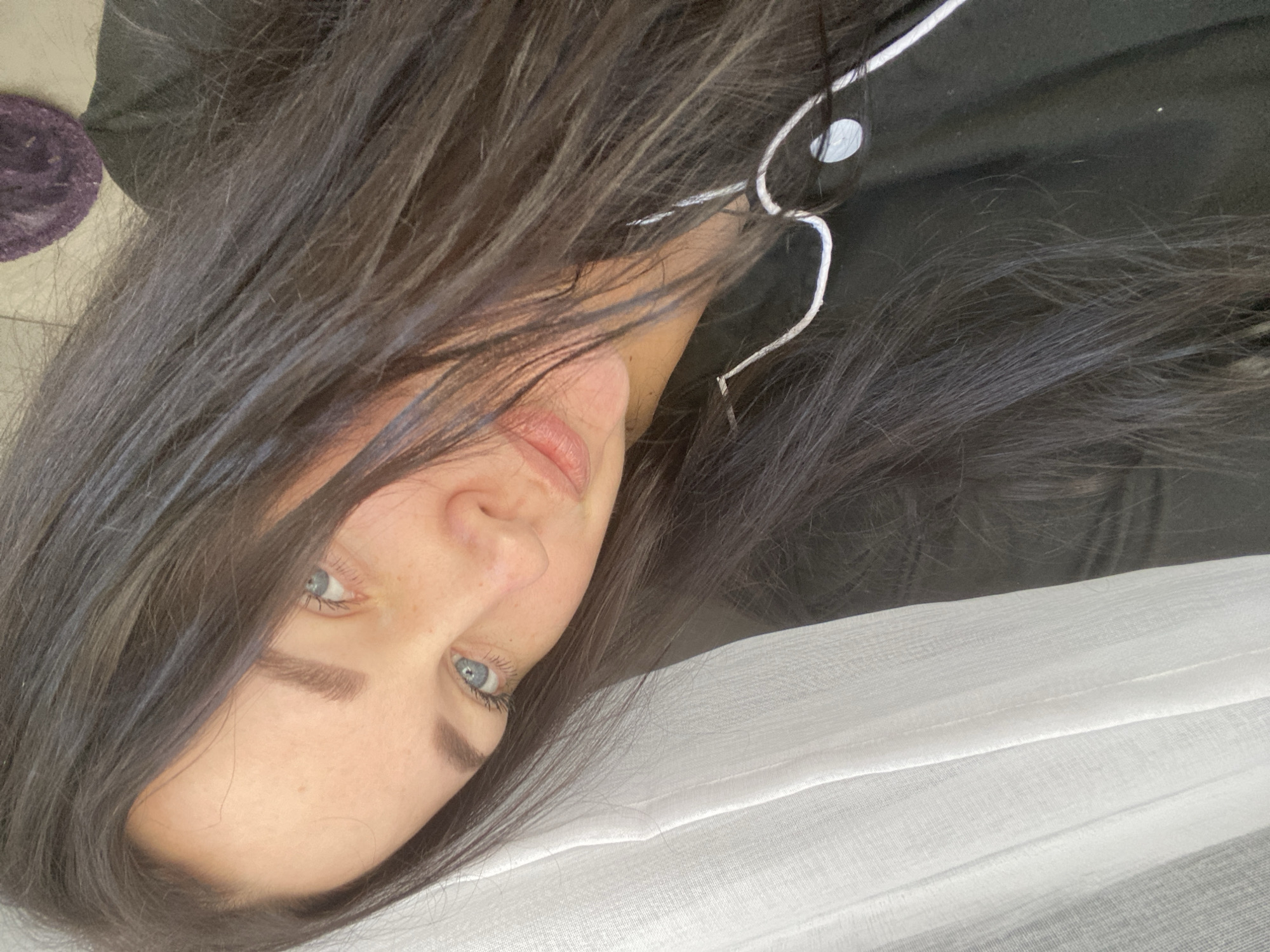 A close-up, tilted portrait features a young adult female with long, dark brown hair, lying down and looking directly at the camera. Her fair-skinned face is partially framed by her disheveled hair, revealing striking blue-grey eyes, dark eyebrows, and natural-looking lips. She exhibits a neutral expression, engaging the viewer with direct eye contact.
She is dressed in a black top detailed with white piping and a white drawstring around the neckline or collar, suggesting casual loungewear or pajamas. The setting appears to be indoors, likely a bedroom or a comfortable living space in Sochi, Russia. To the right and bottom of the frame, a white, subtly textured, possibly sheer fabric is visible, which could be a bedsheet or curtain. A small glimpse of a dark, textured object with a hint of purple is present in the top-left corner, possibly a blanket or cushion. The lighting is soft and diffused, contributing to the relaxed ambiance of the scene. There is no visible text in the image.