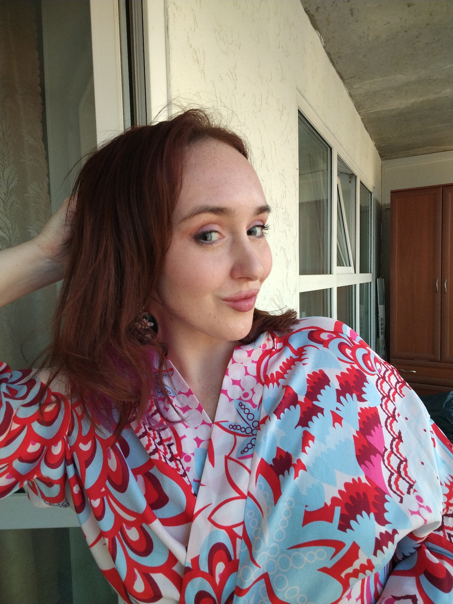 A young woman with reddish-brown hair, fair skin, and light freckles is depicted in a chest-up portrait. She looks towards the viewer with light blue-green eyes and a subtle, pleasant smile, her head tilted slightly. Her left hand is raised, with her fingers gently resting in her hair near the back of her head. She wears a colorful, kimono-style garment adorned with a vibrant abstract pattern featuring shapes in red, blue, pink, and white. A dark, possibly floral or ornate earring is visible on her left ear.
The setting appears to be indoors or on a balcony, illuminated by bright daylight. Behind her, a textured, light-colored wall is visible on the left. To her right, a large window with white frames forms a significant part of the background, with an exposed concrete ceiling above it. A dark wooden cabinet or wardrobe is partially visible on the far right. The overall impression is calm and casual. There is no visible text in the image. The location context provided is Izhevsk, Russia.
