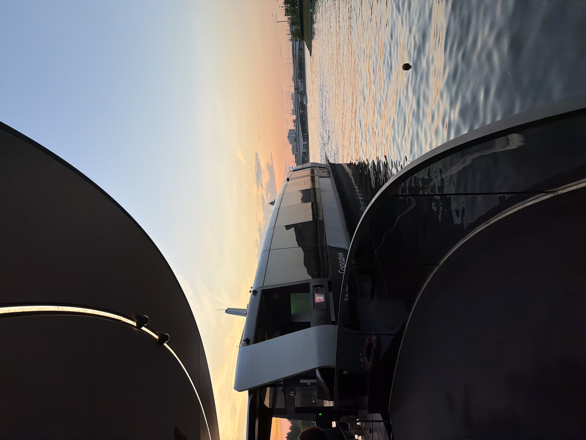 The image depicts a sunset view on a river in Moscow, Russia, with a modern boat docked in the foreground. The boat, which appears to be a passenger vessel, has a sleek, contemporary design with large windows and a dark hull. The word "ГОРОДНЯ" is visible on the side of the boat. In the background, a bridge spans the river, and the city skyline with buildings and lights can be seen. The sky is painted with warm hues of orange and pink, indicative of a sunset. The water is calm, with gentle ripples reflecting the sky's colors. The foreground features a dark, curved structure, possibly part of a pier or a floating platform, with a glass railing. A single, dark, spherical object is floating on the water. The overall scene conveys a sense of tranquility and urban beauty at dusk.
