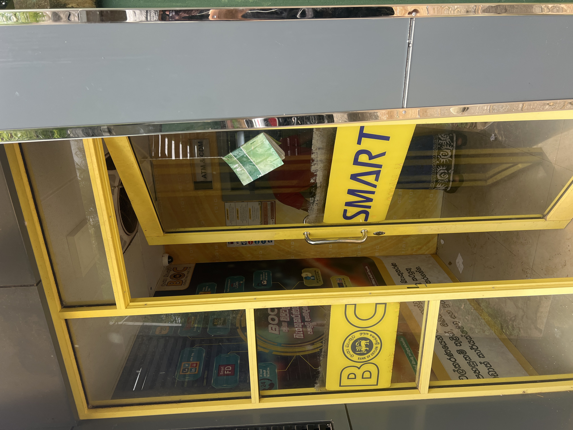 The image shows the exterior of a Bank of Ceylon (BOC) ATM booth in Boossa, Sri Lanka. The ATM booth has a yellow frame and glass doors. One of the glass doors is ajar. Inside the booth, a person wearing a red shirt and a striped skirt can be seen. Various posters and signage related to BOC services are displayed on the walls and doors. A piece of green paper, possibly a withdrawal slip or a note, is hanging from a string inside the booth. The ATM machine itself is visible behind the glass. The exterior wall of the building is gray. The ground appears to be tiled.