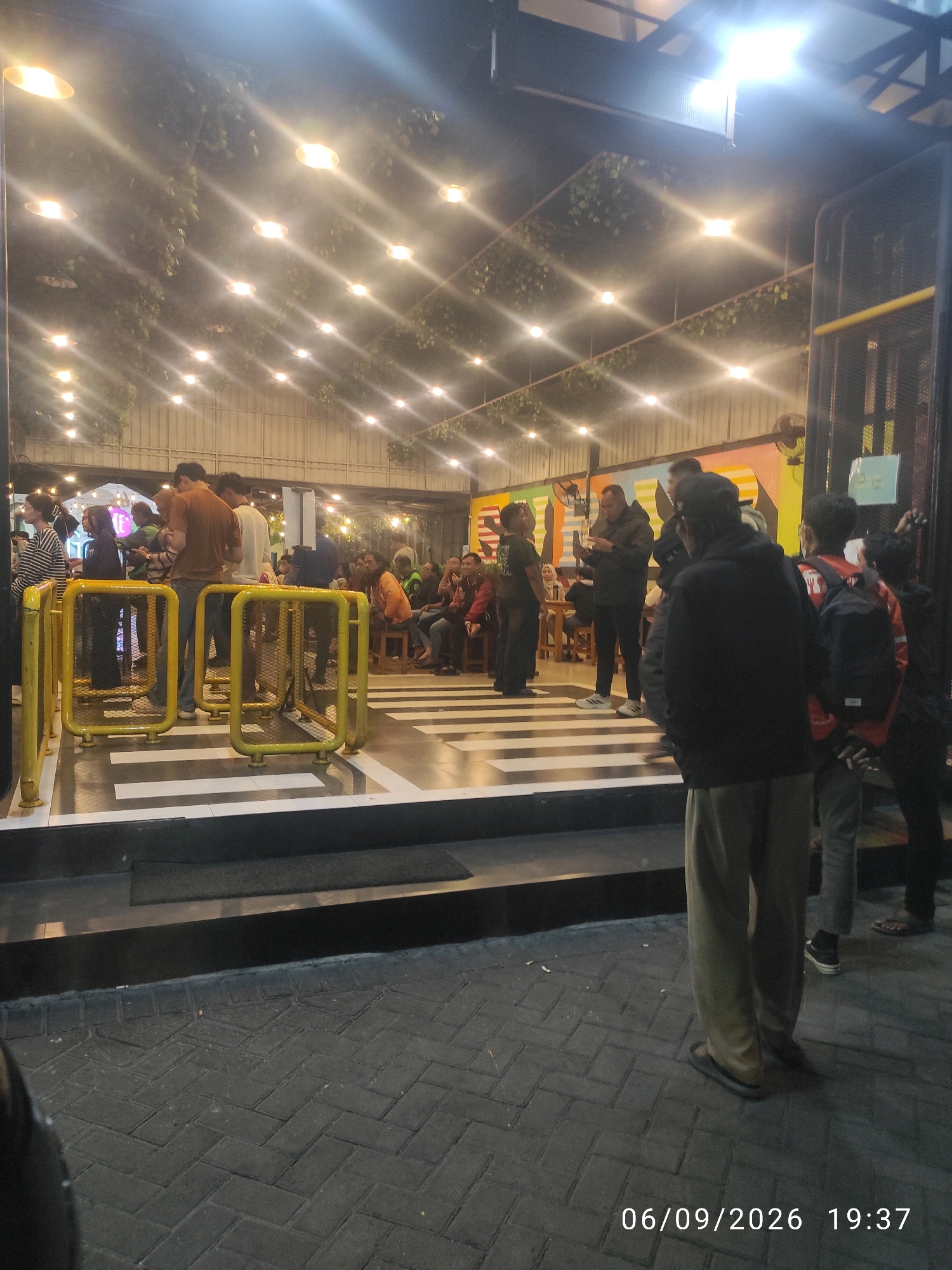 An outdoor nighttime scene depicts a group of people gathered around yellow turnstiles in front of a building with a colorful mural. The building is illuminated by numerous string lights, and greenery is visible hanging from above. In the foreground, a person wearing light-colored pants and flip-flops stands facing away from the camera, with their legs and feet visible. The ground is paved with dark gray interlocking bricks. A timestamp in the lower right corner reads "06/09/2026 19:37". The location is identified as Surabaya, Indonesia.