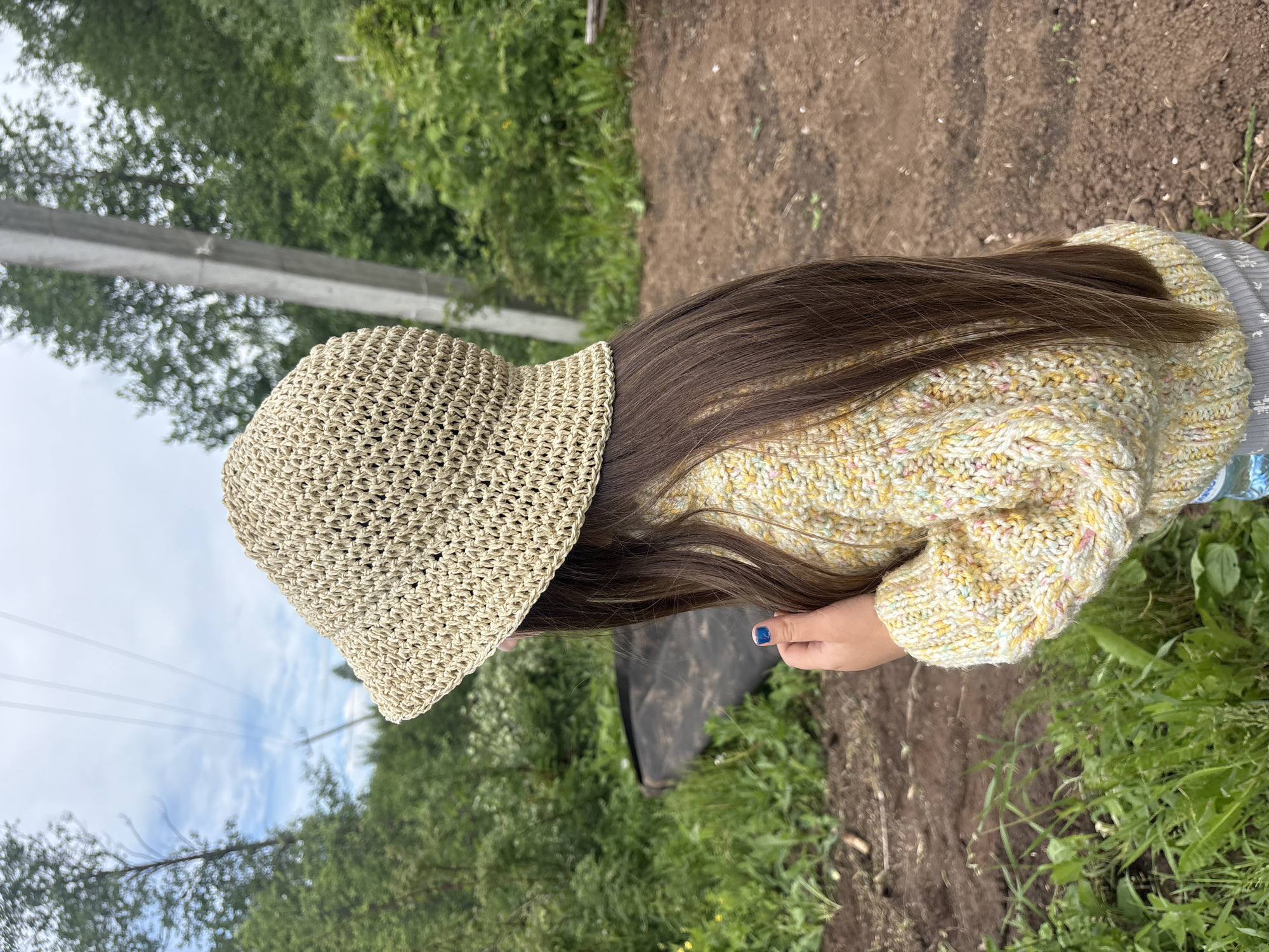 An overhead shot captures a person, likely a child or young individual, viewed from behind, standing outdoors in what appears to be a rural or semi-rural setting in Фоки, Russia. The individual has long, straight, dark brown hair and is wearing a light beige, open-weave crocheted bucket hat. They are dressed in a chunky, knitted sweater or cardigan, primarily yellow with colorful flecks of pink, blue, and white yarn, suggesting a cozy garment. One hand is visible, featuring a fingernail painted with bright blue nail polish.
The ground beneath the person is a mix of dark brown, earthy soil on the right side of the frame and vibrant green grass and weeds on the left. In the mid-ground and background, a dense expanse of green trees and foliage creates a natural backdrop. Above the treeline, a soft, overcast sky with visible clouds suggests a cloudy day. A utility pole with several power lines extends across the middle distance, adding a subtle element of infrastructure to the natural landscape. The image conveys a serene outdoor moment during daylight hours. No visible text is present in the scene.