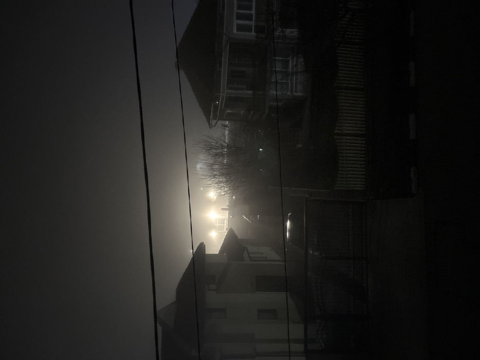 The scene depicts a residential area in Stefan Karadja, Bulgaria, at night during heavy fog. Two prominent dark power lines span horizontally across the upper and middle sections of the frame. On the left, a multi-story house with a light-colored facade and a dark gabled roof is partially visible, featuring a window and a dark entrance. A dark, possibly metal, ornate gate is closed below this house, obscuring a driveway where a dark vehicle's roofline is faintly discernable through the mist. On the right, another multi-story house with a dark, gabled roof, a veranda, and multiple visible windows is present. A wooden picket fence with a scalloped top edge runs along the lower portion of the right side. Between the houses, a bare-branched tree or shrub stands. In the distant background, several intense artificial lights pierce through the dense fog, creating a diffused, bright glow that obscures any specific structures beyond them. No individuals are visible, and no actions or interactions are taking place.