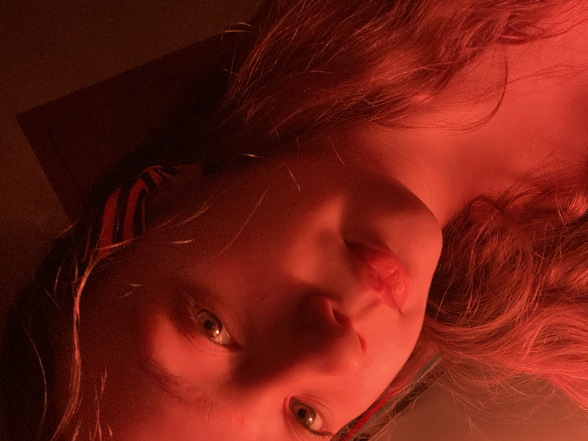 A close-up, canted, frontal view captures a fair-skinned individual with reddish-brown hair. The person's face is prominently lit in a red hue, emphasizing the features: eyes, nose, mouth, and cheekbones. The eyes are light-colored with dark pupils. The person's head is angled, and the image is taken from a slightly lower vantage point. The background is dim and indistinct, with a dark, flat object visible in the periphery. The location and context are unknown.