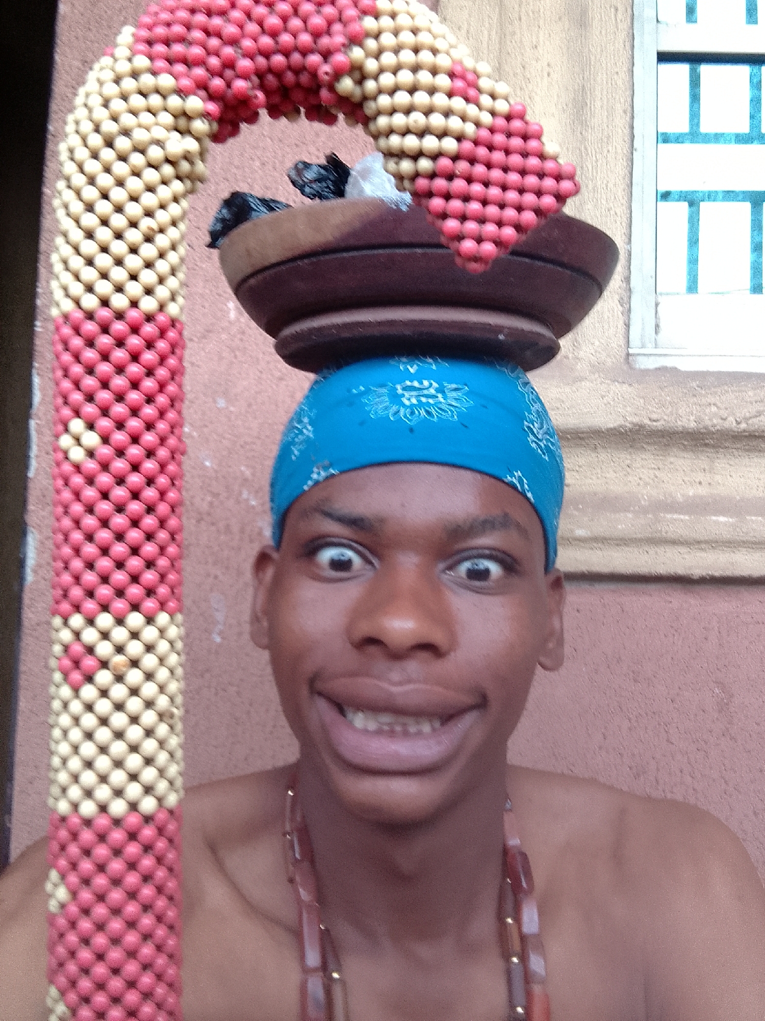 A person with a blue bandana on their head is balancing a wooden bowl on their head. The bowl contains dark items, possibly wrapped in plastic. An arch-shaped beaded structure, decorated with alternating sections of red and off-white beads, is positioned beside the person. The person has wide eyes and a toothy smile. They are wearing a beaded necklace and appear to be standing in front of a wall with a window visible in the background. The city and specific location are unknown.