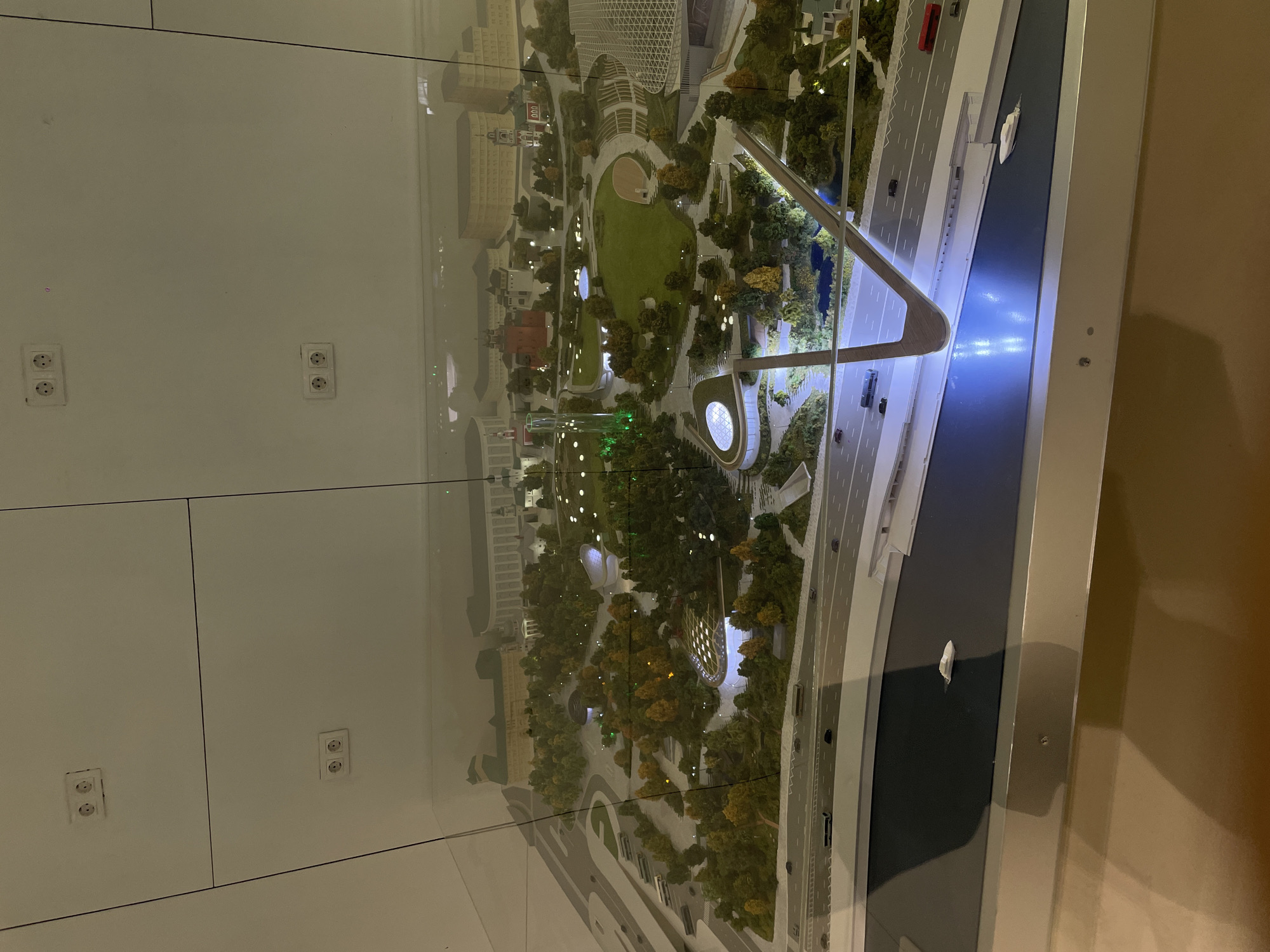 This image shows a detailed architectural model of a cityscape, likely located in Khimki, Russia, viewed through a glass display case. The model features a central park area with a green lawn, trees, and walkways. There are also various buildings depicted, including a large concert hall with a grid-like roof structure, and other structures reminiscent of residential or commercial buildings. A prominent element is a modern, arch-shaped bridge spanning a simulated river or waterway. Miniature vehicles are visible on the roads, indicating the scale and level of detail in the model. The environment includes representations of trees and landscaping. The model is illuminated, casting light on the scene. The background consists of a plain wall with electrical outlets, suggesting that the display is located indoors, possibly in a showroom or exhibition space.
