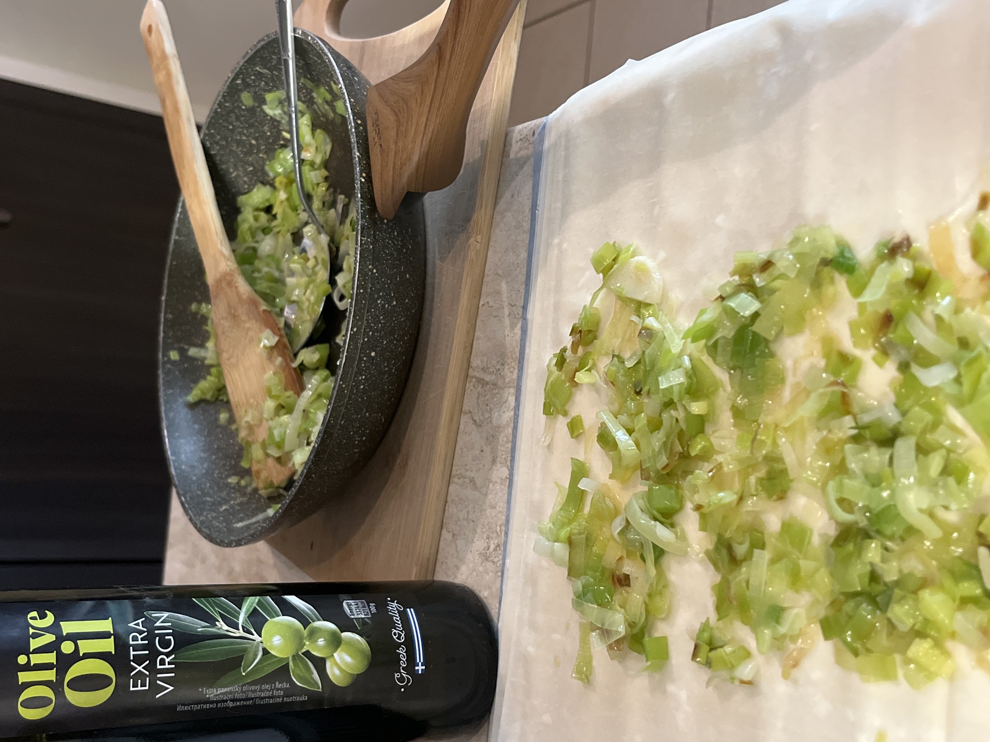 The image shows a cooking scene, likely in a domestic kitchen. The dominant elements are a black frying pan containing chopped leeks being stirred with a wooden spoon and a silver spoon, a baking sheet or parchment paper with more of the same chopped leeks spread on it, and a bottle of "Olive Oil EXTRA VIRGIN," which indicates the olive oil is of Greek origin. The bottle is black with a label featuring green text and images of olives. No people are visible in the frame. The setting implies food preparation in progress. There is no information available about the location of Sofia, Bulgaria.