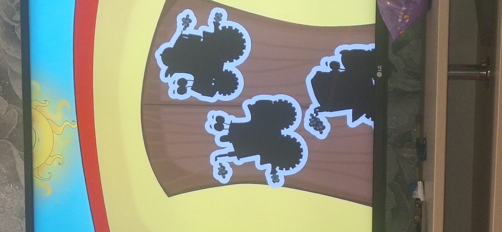 This is a television screen displaying a cartoon image with a cartoon sun and rainbow in the upper portion. The main focus is on three black tractor silhouettes against a brown, wood-like background. The tractors have white outlines. One tractor is on the left, one is in the upper right, and the third is in the lower center. The image does not contain any identifiable elements related to Bila Tserkva, Ukraine. The screen is positioned above a shelf with various items, including a pair of white and blue shoes, and a purple bag. An LG logo is visible on the bottom bezel of the television.