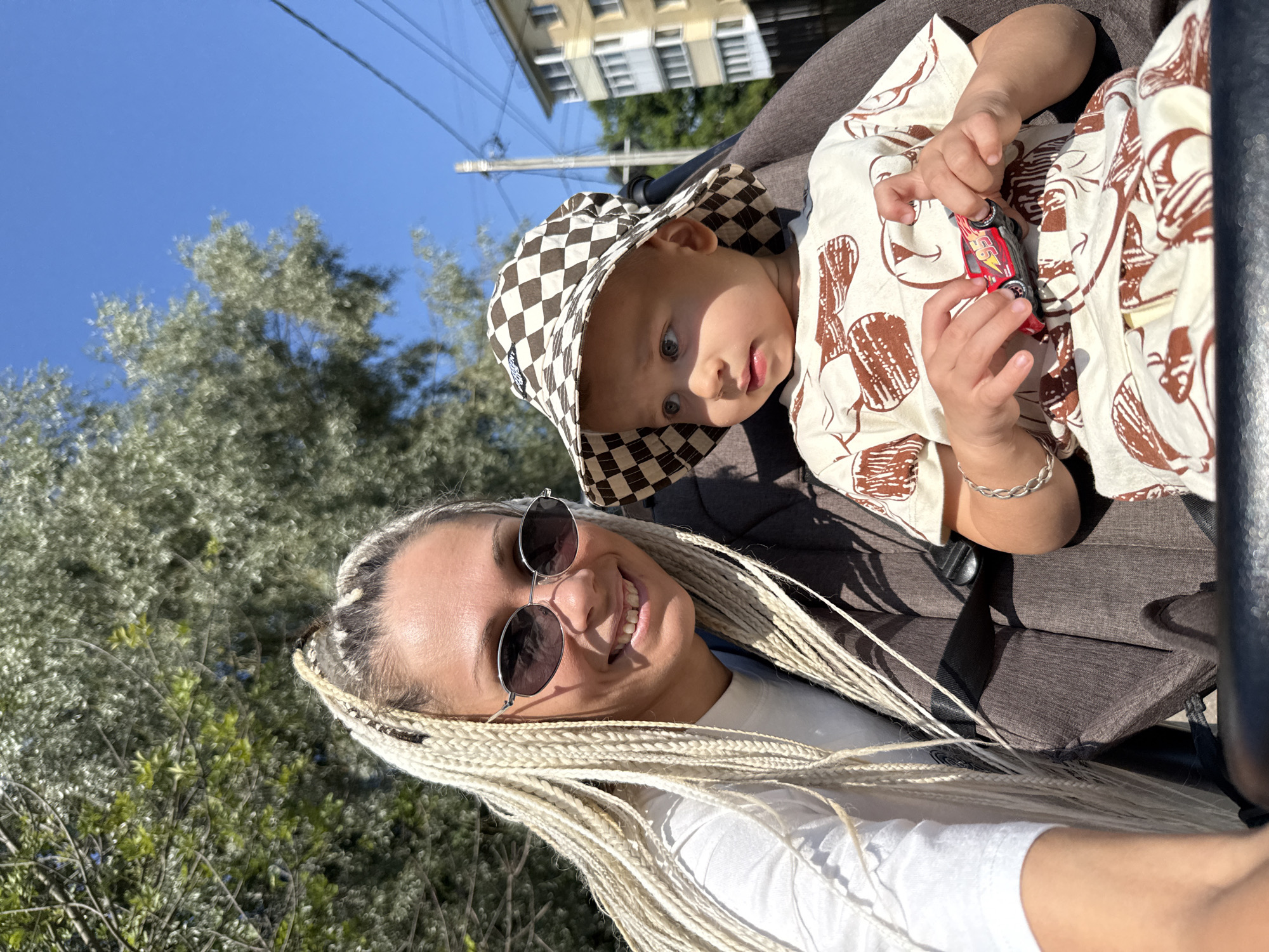 A selfie taken outdoors shows a woman with long blonde braided hair and sunglasses smiling, with a young child in a stroller in the foreground. The woman is wearing a white t-shirt. The child is wearing a checkered bucket hat and a light-colored t-shirt with a brown pattern. The child is holding a red toy car. The background includes a large tree with green leaves and a clear blue sky. In the distance, there is a multi-story building and a utility pole with wires. The scene suggests a sunny day in a residential area.