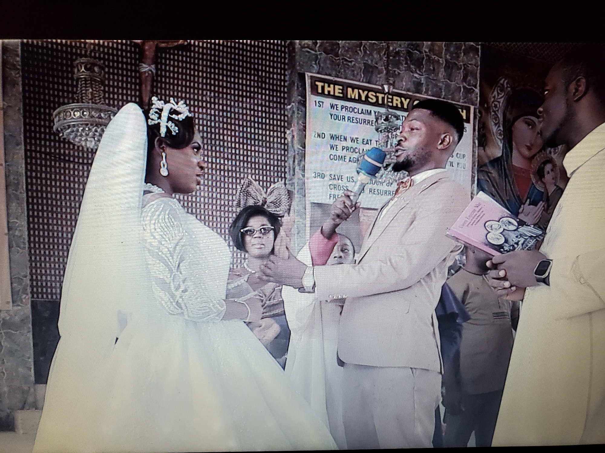 A wedding ceremony appears to be in progress indoors, possibly in a church setting. The bride, wearing a white wedding dress, veil, and tiara, faces a man speaking into a microphone. He is dressed in a light-colored suit and holds a pink book in his left hand. Another man is standing to his right in a white shirt. Behind the couple, a woman with glasses is visible, and a sign reads "THE MYSTERY." A religious painting is visible on the wall near the man in the white shirt, and a crucifix hangs on the wall behind the bride.
