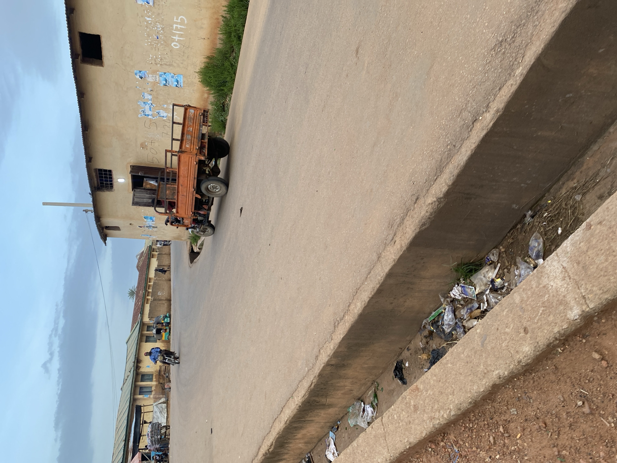 This is a medium shot, eye-level, exterior photograph of a street scene in Bukuru, Nigeria. The image captures a dirt road with a concrete drainage ditch running along the left side. The ditch is filled with refuse, including plastic bags, bottles, and other debris. On the right side of the road, a light-colored building with a corrugated metal roof is visible. The building has several open windows and posters affixed to its facade. An orange three-wheeled truck with a cargo bed is parked on the road in front of the building. The truck has a single driver visible in the cab. Further down the street, a person is riding a motorcycle. In the background, there are more buildings, some with awnings, suggesting a commercial or residential area. The sky above is overcast with clouds. The overall impression is that of a dusty, somewhat cluttered urban environment.