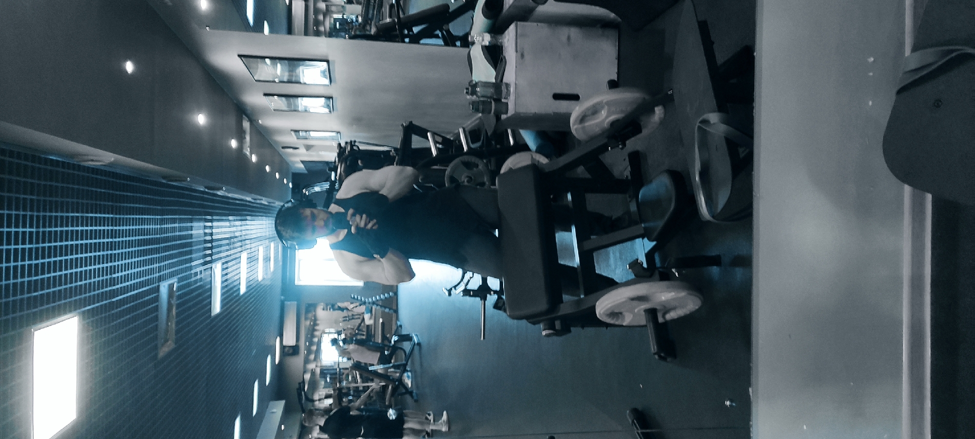 The image depicts a person wearing a black tank top and headphones standing in what appears to be a gym in Fryazino, Russia. The person is positioned near exercise equipment, potentially working out. Other gym equipment and possibly other individuals can be seen in the background. The gym has a modern aesthetic with visible lighting fixtures and a patterned wall.