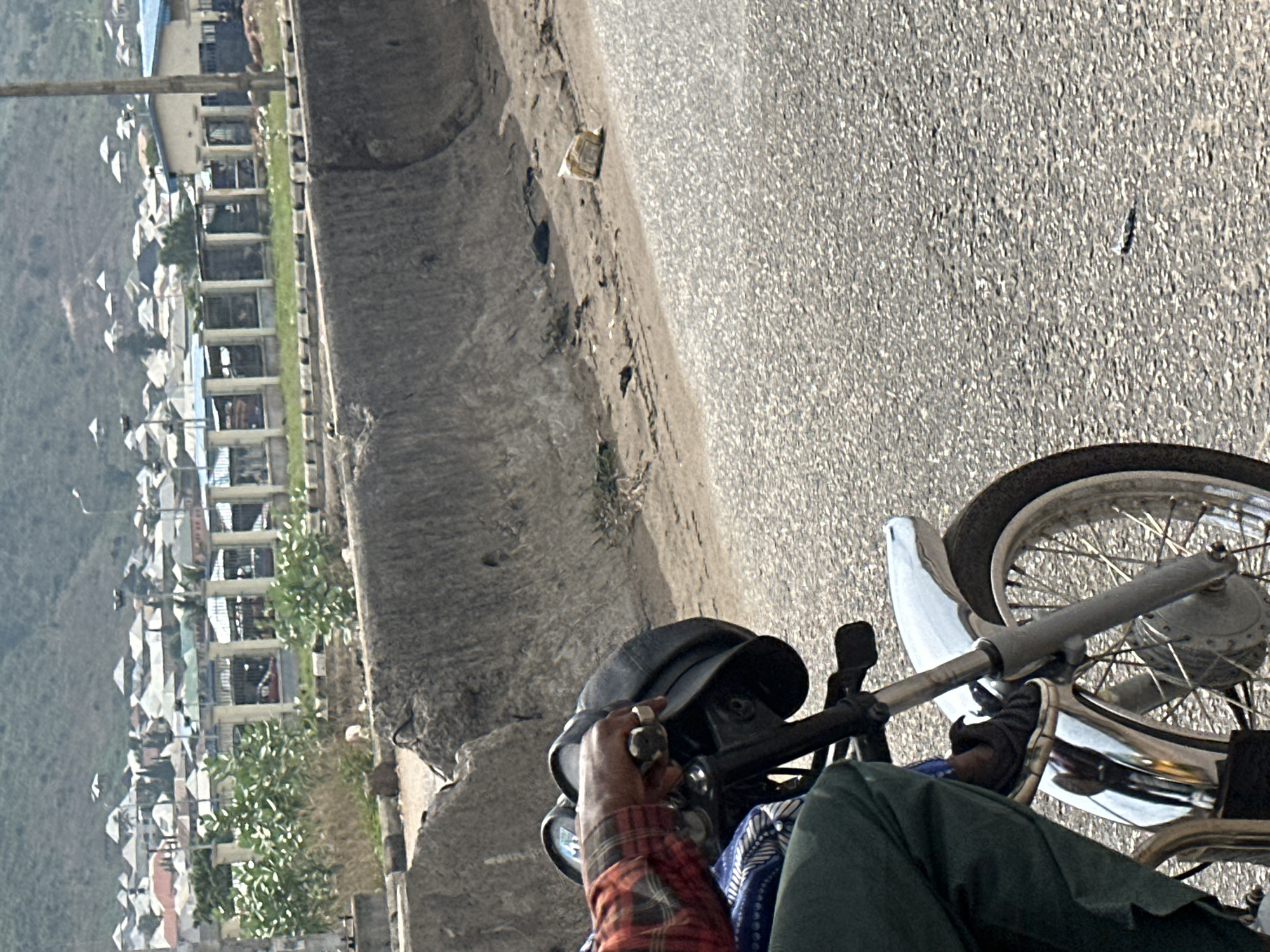 The image shows a first-person perspective of someone riding a motorcycle on a paved road, likely in Lokoja, Nigeria, based on the architecture in the background. The motorcycle's front wheel and handlebars are visible. The rider is wearing a cap, a red plaid shirt, and green pants. A concrete curb and debris are along the roadside. In the distance, there are buildings with distinctive white roofs. Vegetation is visible interspersed with the buildings.