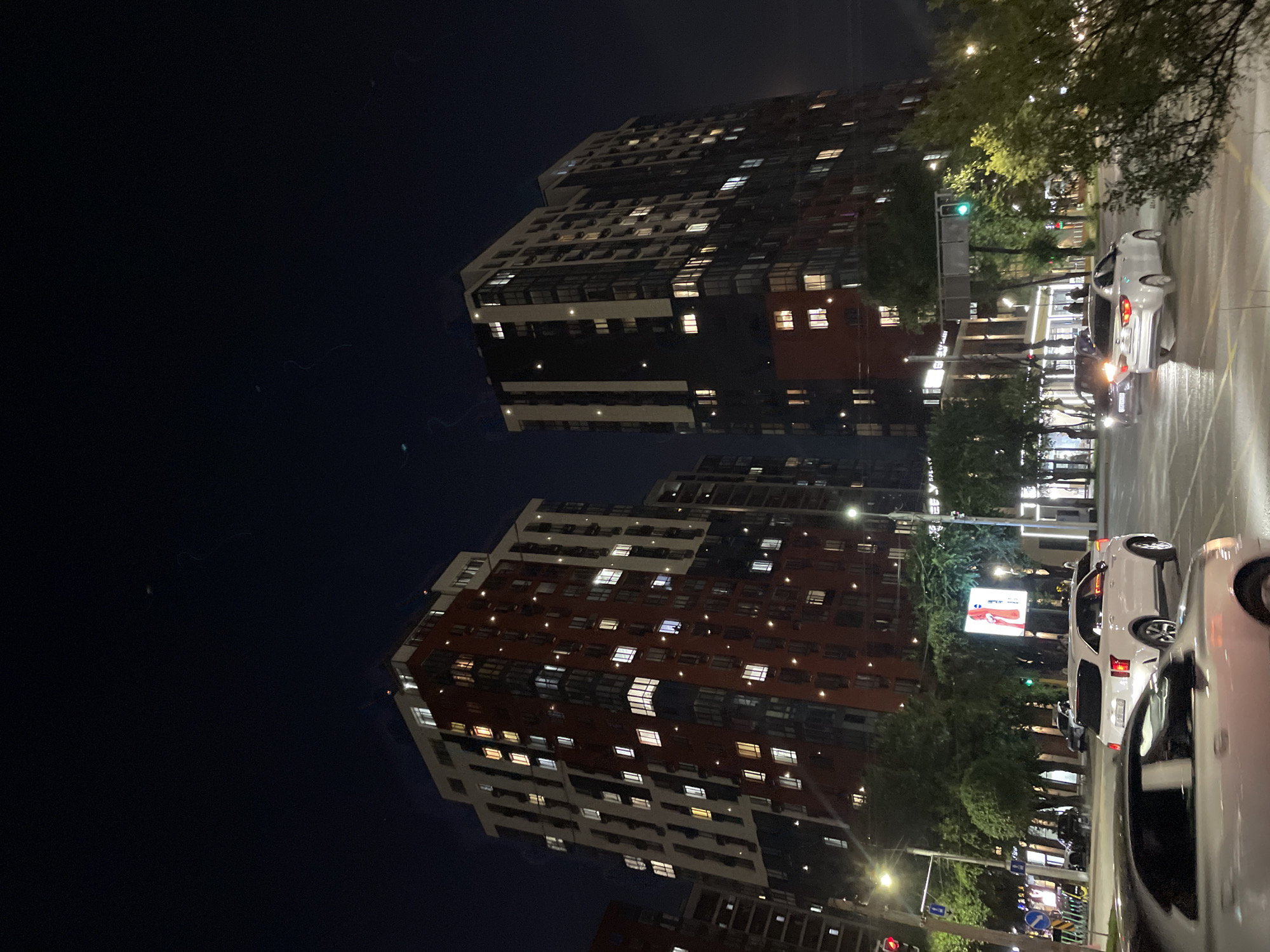 This is a night-time street-level view in Almaty, Kazakhstan, with two tall, modern apartment buildings dominating the background. The buildings are illuminated by numerous lit windows, indicating occupancy. The left building is predominantly red and white, while the right building is dark gray and white. The street is wet, suggesting recent rain, and several cars are visible, including a white SUV and a silver sedan in the foreground. Traffic lights are visible, with one showing green. Trees line the street, and some smaller buildings and businesses are present at the base of the apartment complexes. The sky is dark, with some faint light streaks that could be lens flares or atmospheric phenomena.
