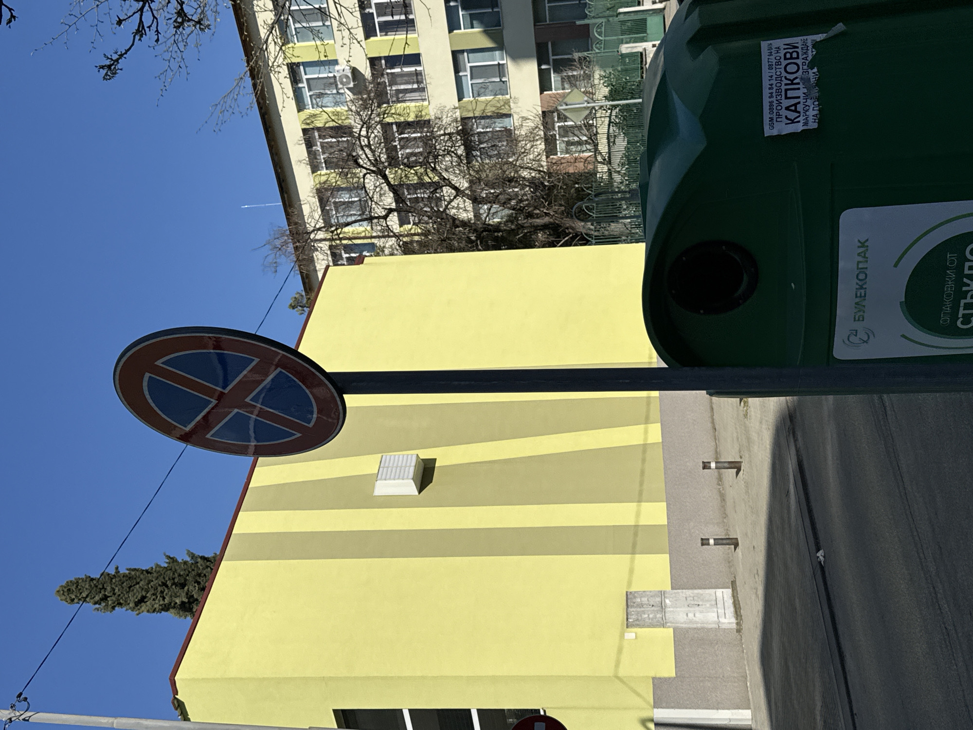 Here's a detailed description of the image:
**Scene Overview:** The image presents an outdoor urban scene, likely in Stara Zagora, Bulgaria. The view is angled upwards, capturing elements of street infrastructure, buildings, and environmental features under a clear blue sky.
**Objects:**
* **Traffic Sign:** A "No Parking" sign is prominently displayed.
* **Buildings:** There are two buildings visible. One has a façade with horizontal yellow and light-yellow stripes. The second building appears to be an apartment building with many windows.
* **Waste Container:** A large green waste container is on the right side of the image. It features labels indicating its use for glass recycling, including the Bulgarian company "Булекопак" and contact information for a company specializing in drip irrigation systems.
* **Street/Sidewalk:** A portion of the street or sidewalk is visible to the right of the yellow building, with street elements like metal rods.
* **Vegetation:** A tree with bare branches is near the buildings. A tall conifer is seen near the building with the traffic sign.
**Environment:**
* **Sky:** A clear, blue sky provides the backdrop.
* **Weather:** The lighting suggests it's a sunny day.
**Specific Details:**
* The label on the waste container indicating glass recycling.
* The architectural design of the buildings.
* The presence of a "No Parking" traffic sign.
The image provides information about waste management, urban environment, and traffic control in Stara Zagora.
