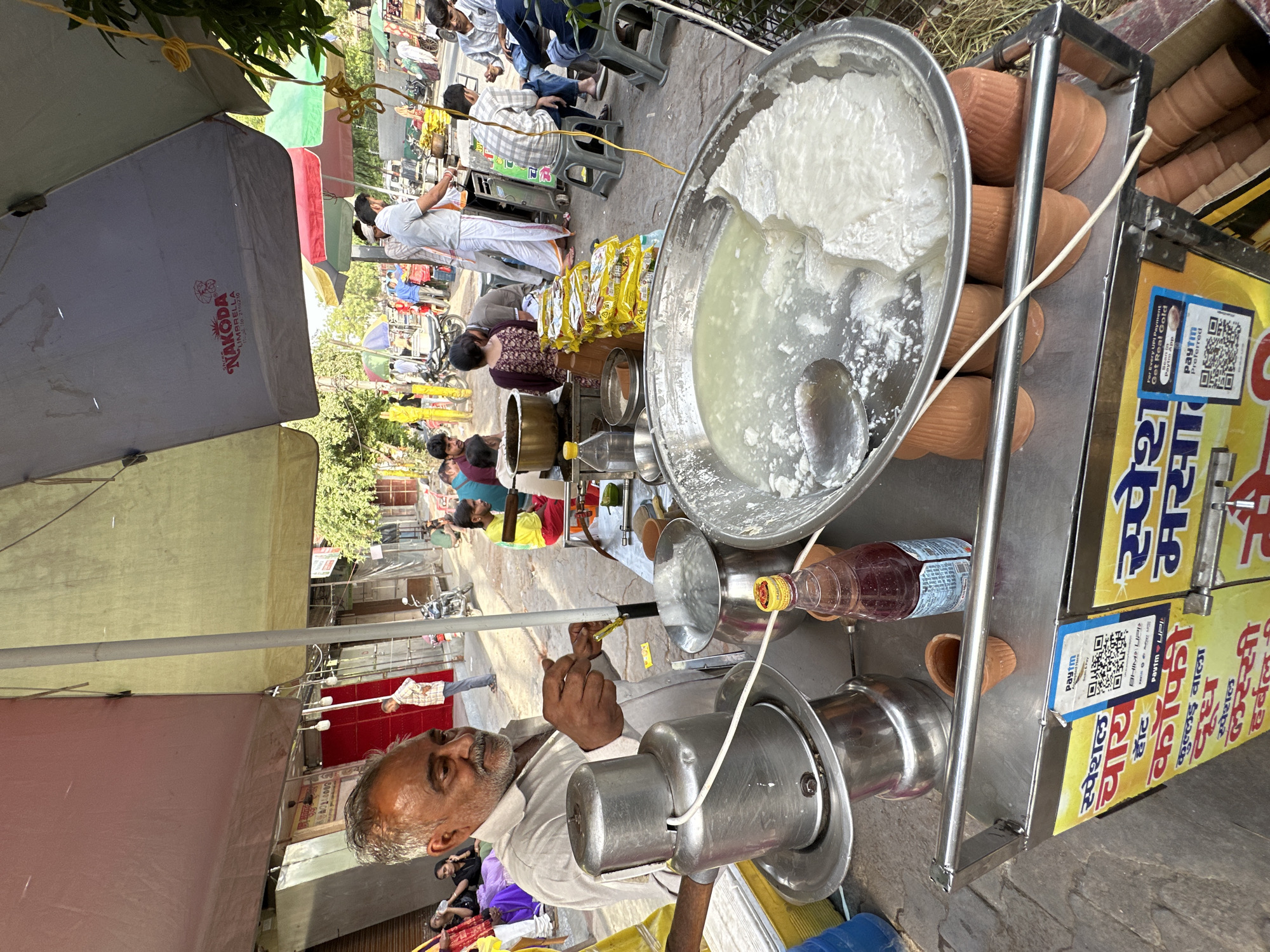 Here's a detailed description of the image:
The image shows a street scene in Vrindavan, India. An Indian man, likely a street vendor, is operating a food cart. He is standing under a "NAKODA Umbrella" brand umbrella, indicating the cart is in a semi-permanent location.
The cart contains a variety of items related to the preparation and sale of what appears to be a milk-based drink or dessert. A large silver bowl contains a white, creamy substance, possibly curd or yogurt, partially mixed with liquid. There's also a metal container with what seems to be a beverage-making machine, probably for lassi. Several clay cups (kulhads) are stacked next to the bowl, indicating individual servings are prepared and sold. A bottle of red syrup is also on the cart. A sign in Hindi script is attached to the cart, likely advertising the items for sale, along with a QR code for digital payment (Paytm).
In the background, a street scene is visible with people walking by, suggesting a busy location. Some individuals are seated. There are various shops and stalls with colorful umbrellas, typical of Indian marketplaces.