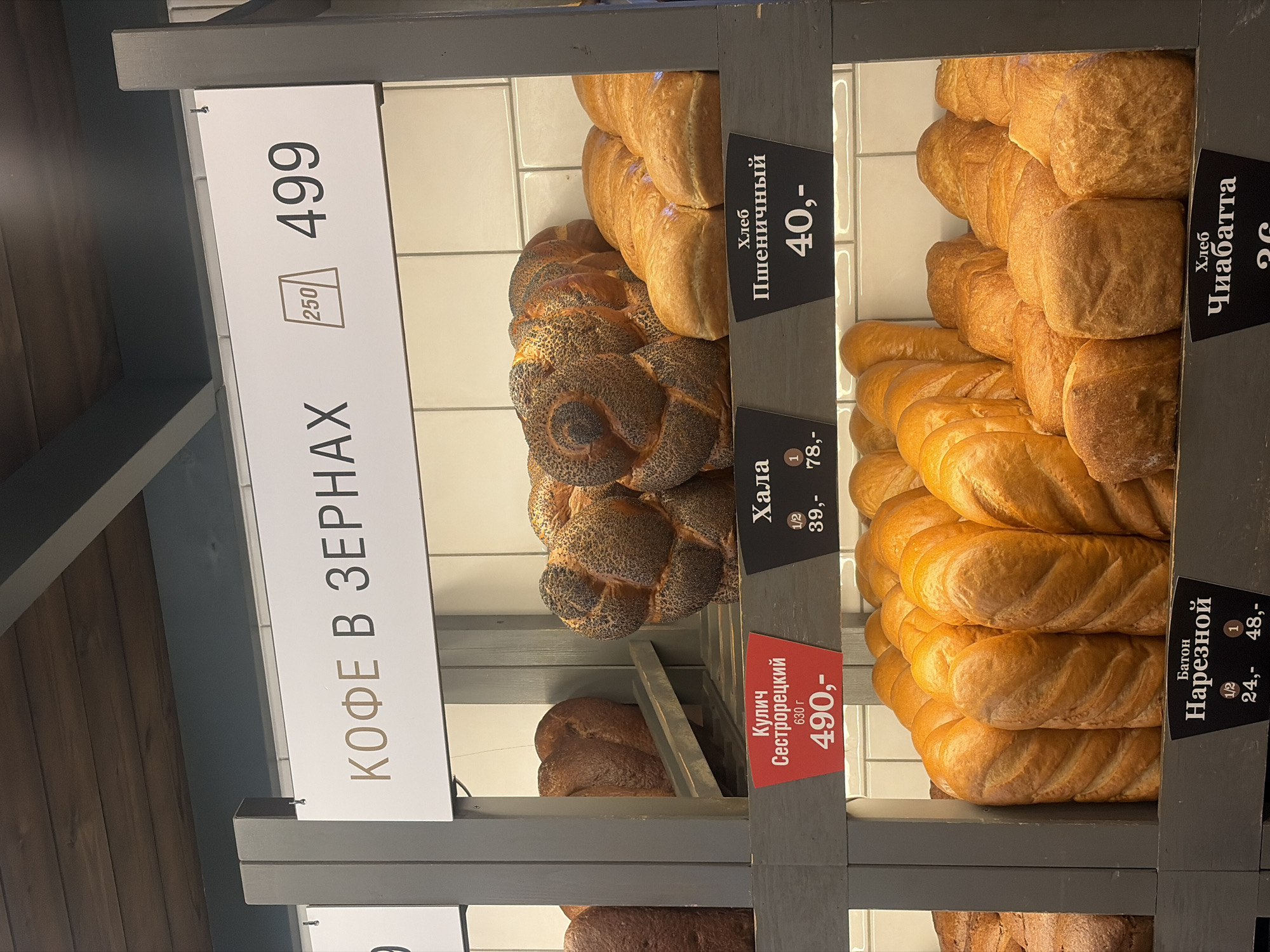 This is an indoor shot of a bread display in a shop, probably in Saint Petersburg, Russia, given the Cyrillic alphabet. The display features various kinds of bread, including 'Khala,' 'Sestroretsky Kulich', 'Wheat Bread' and sliced baton loaf ('Nareznoy Baton'). There is also a sign advertising 'Coffee Beans' (Kofe V Zernakh) at a price of 499. Price tags are visible for each type of bread, ranging from 24 to 78. The shop has white tiled walls behind the display. The bread loaves are stacked neatly.