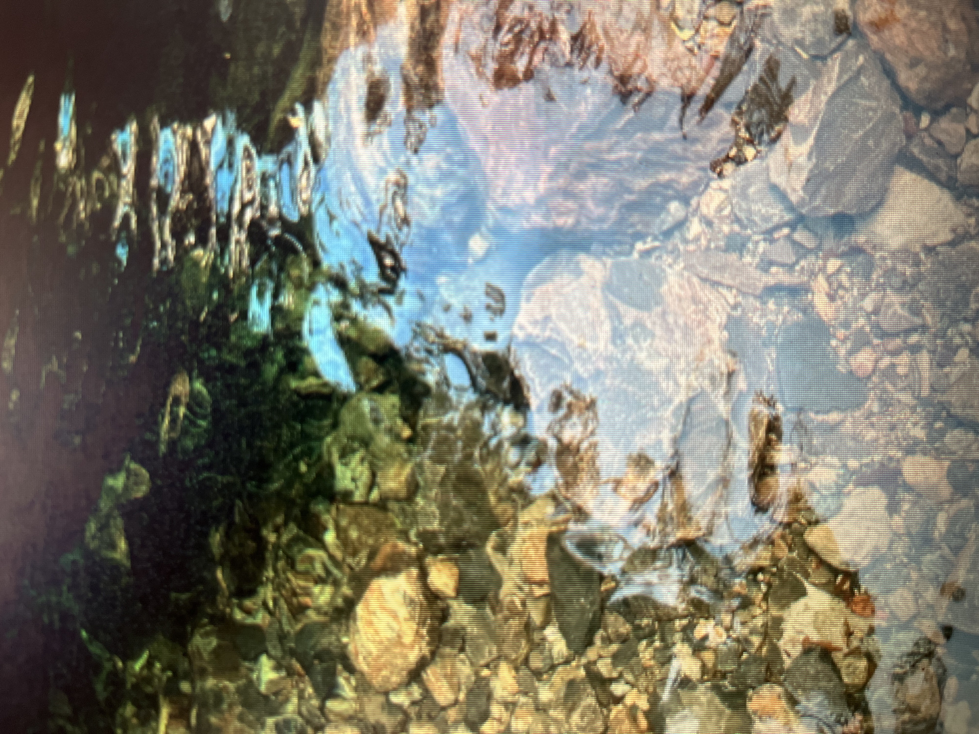 The image shows a shallow, rocky stream bed with clear water. The rocks are of various sizes and colors, including beige, brown, and gray. Some submerged aquatic plants are visible, adding a dark green hue to the scene in some areas. The water's surface reflects the sky, creating patches of bright blue and white light that distort the view of the rocks and plants below. The overall impression is one of a natural, undisturbed aquatic environment. The location is not specified as being in the city of Sejahtera, Indonesia.