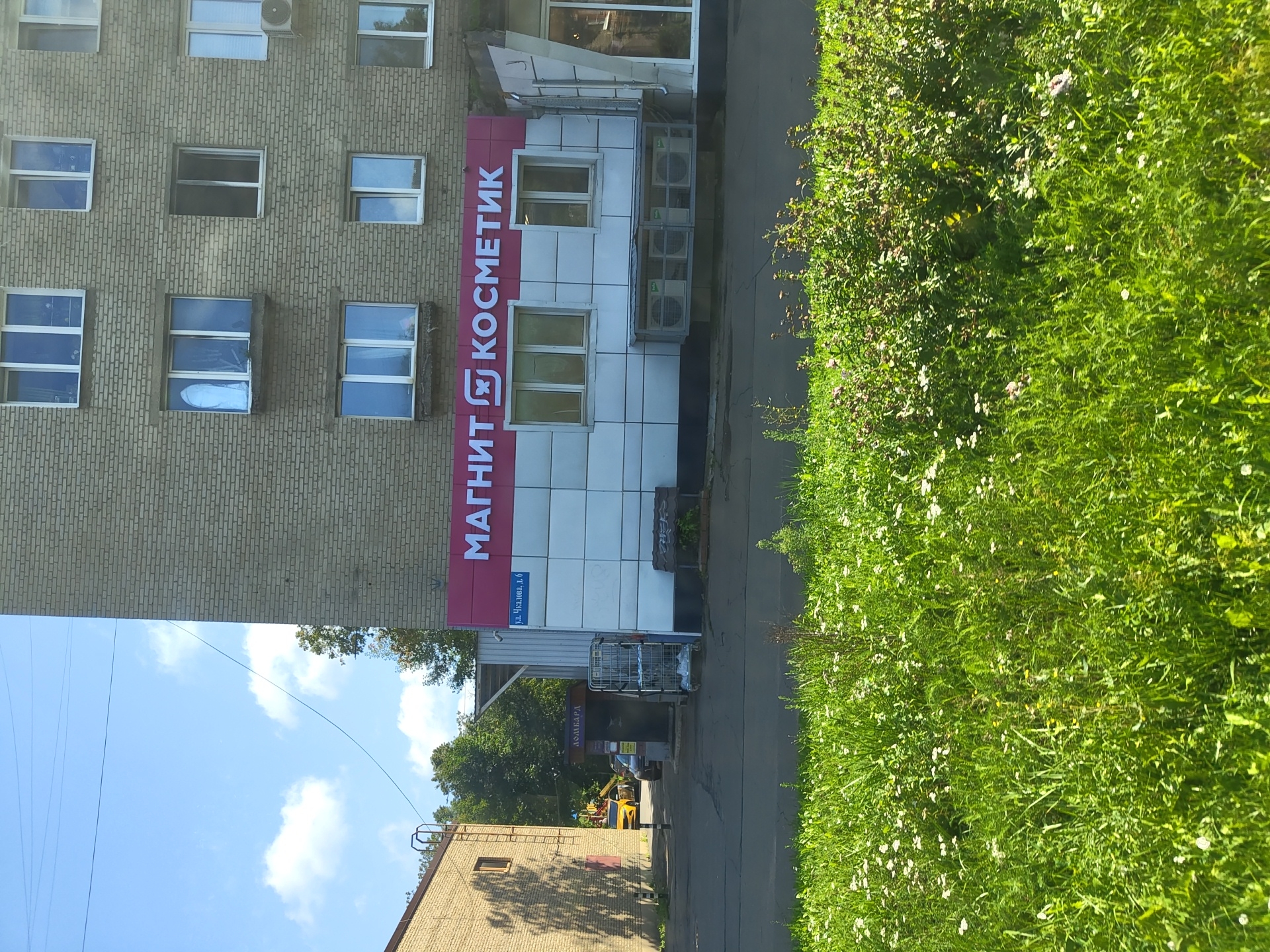 The image shows the storefront of a "Magnit Kosmetik" store located in the city of Zhukovsky, Russia. The store is situated on the ground floor of a multi-story brick building. The storefront features a large, prominent sign with the store's name in white lettering on a pink background. Below the main sign, there is a smaller blue sign that reads "ул. Чкалова, д. 6", indicating the street address is Chkalova Street, building number 6. The building itself has multiple windows on its upper floors. In front of the store, there are several air conditioning units installed under a sheltered area. To the left of the store, there is another building, and a yellow car is visible further down the street. The area in front of the buildings appears to be a paved walkway or street, with a grassy area in the foreground. The sky is blue with some clouds, suggesting it is daytime.