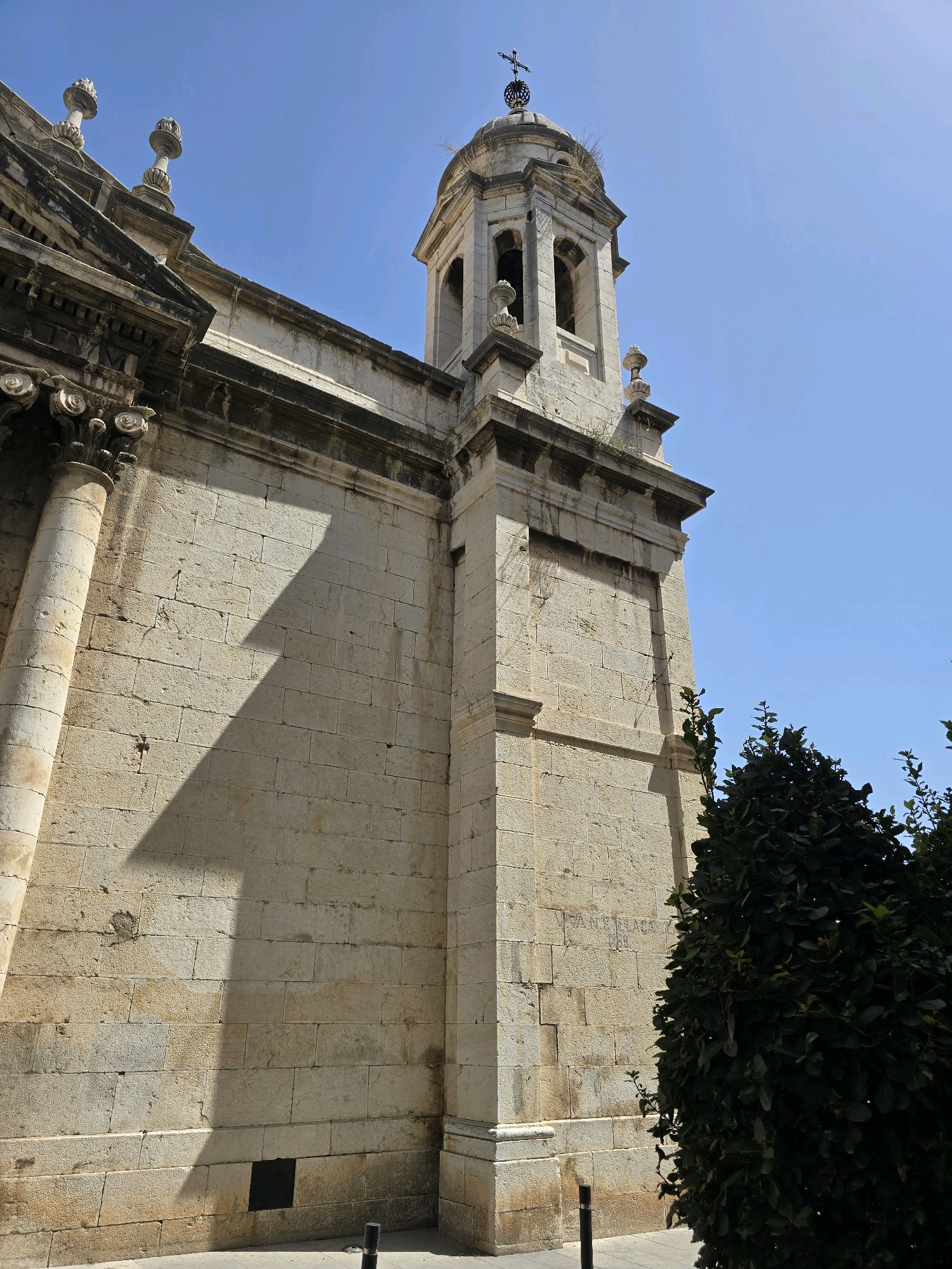 The image shows the side of a stone building with a tower, likely a church, under a clear blue sky. The building is constructed of large, rectangular stone blocks, showing signs of age and weathering. A prominent feature is the bell tower, which is topped with a dome and a cross. The architecture appears to be of a classical or baroque style, with decorative elements visible on the building's facade, including columns and cornices.
A large, dark shadow of a triangular shape is cast across the building's wall, indicating the presence of an object or structure casting the shadow and the sun's position. To the right of the building, a dense, dark green shrubbery or tree is partially visible, obscuring some of the building's lower sections. The ground in front of the building appears to be paved, with the base of the building meeting the pavement. There are two dark bollards on the pavement near the base of the building.
The building's facade has some inscribed markings, partially visible on the stone blocks. Based on the architectural style and typical landmarks in Jaén, Spain, this building is likely the Church of Santa María Magdalena. This church is located in the historic center of Jaén.