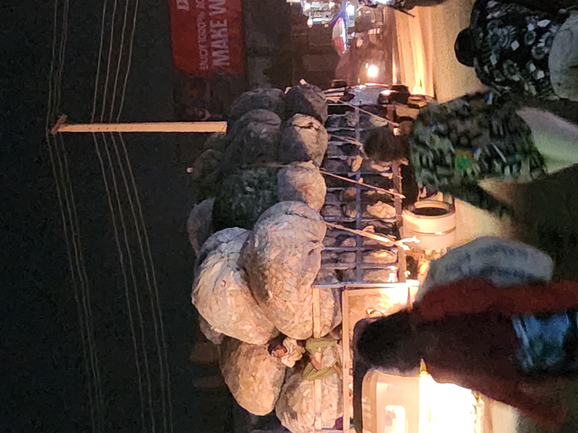 Here is a detailed description of the image:
The image depicts a street scene at night, likely a market or roadside vendor setup in an urban area in Unknown, Unknown. The composition is somewhat tilted. Several large sacks or bundles are stacked high on a metal rack or cart, likely goods being transported or sold. Overhead electrical wires are visible on the left side of the image.
Several people are present. Some appear to be customers or passersby. One person in the foreground is wearing a red shirt and interacting with something or someone near the cart. Other individuals are wearing patterned shirts.
The overall environment is illuminated by streetlights and vendor lights, creating bright spots against the dark backdrop. There is a sign with partially visible text, "ENJOY 100% AC... MAKE W..." which indicates a commercial establishment.