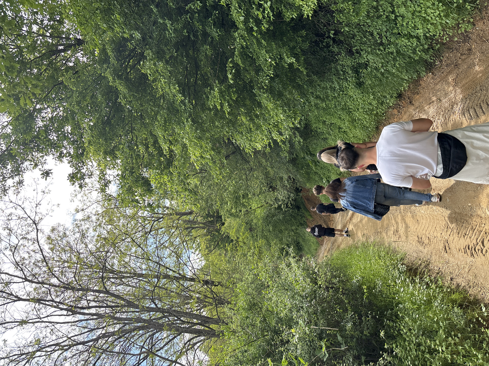 People are walking along a dirt path surrounded by lush greenery, likely in a wooded area. The path shows signs of tire tracks. A person wearing a white shirt and light-colored pants is prominent in the foreground. Other individuals are visible further down the path, one wearing a denim jacket and another wearing a black outfit. There are trees with dense foliage. Given the prompt constraints, the image is suggestive of a recreational outdoor activity, potentially hiking or walking in the vicinity of Zlatograd, Bulgaria, although specific landmarks confirming the location are absent.