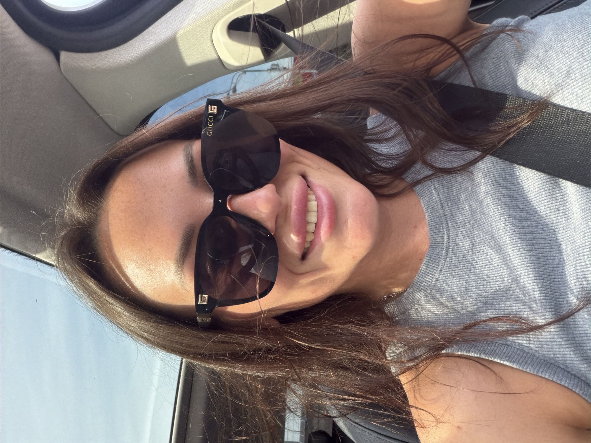 The image shows a woman inside a car, possibly driving, as indicated by the seatbelt and the background visible through the windshield. She is wearing large, dark-rimmed sunglasses with the "GUCCI" logo visible on the side. The woman has long, dark brown hair and is smiling, revealing her teeth. She is wearing a grey ribbed tank top and a delicate silver necklace. The lighting suggests it is daytime and sunny. The background appears to be a city environment with some buildings and streetlights visible, but no specific landmarks of Moscow are identifiable.