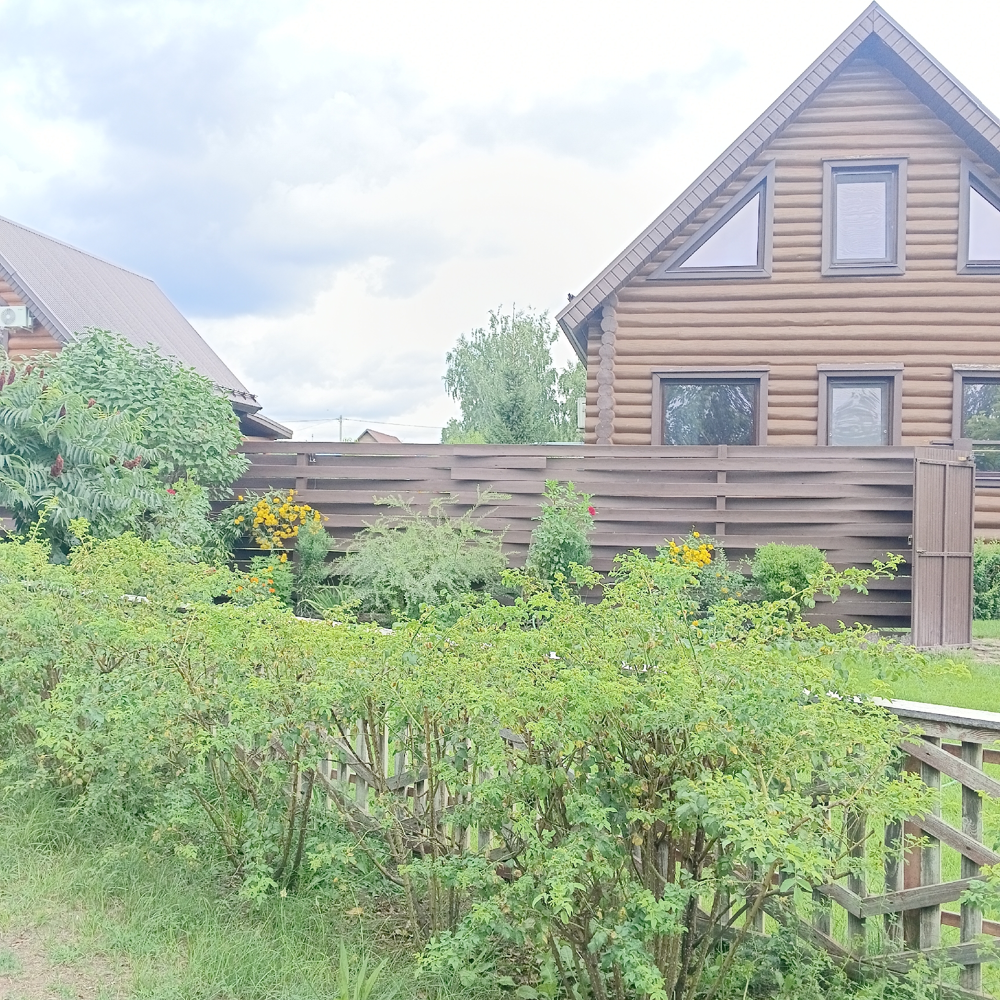 The image depicts a wooden log house in the city of Rozhdestveno, Russia, with a dark wooden fence in the foreground and lush green bushes and trees. The house has a brown metal roof and several windows, including triangular ones on the upper level. The fence is made of horizontal wooden planks. Yellow flowers and other greenery are visible behind the fence. The sky is overcast with clouds.